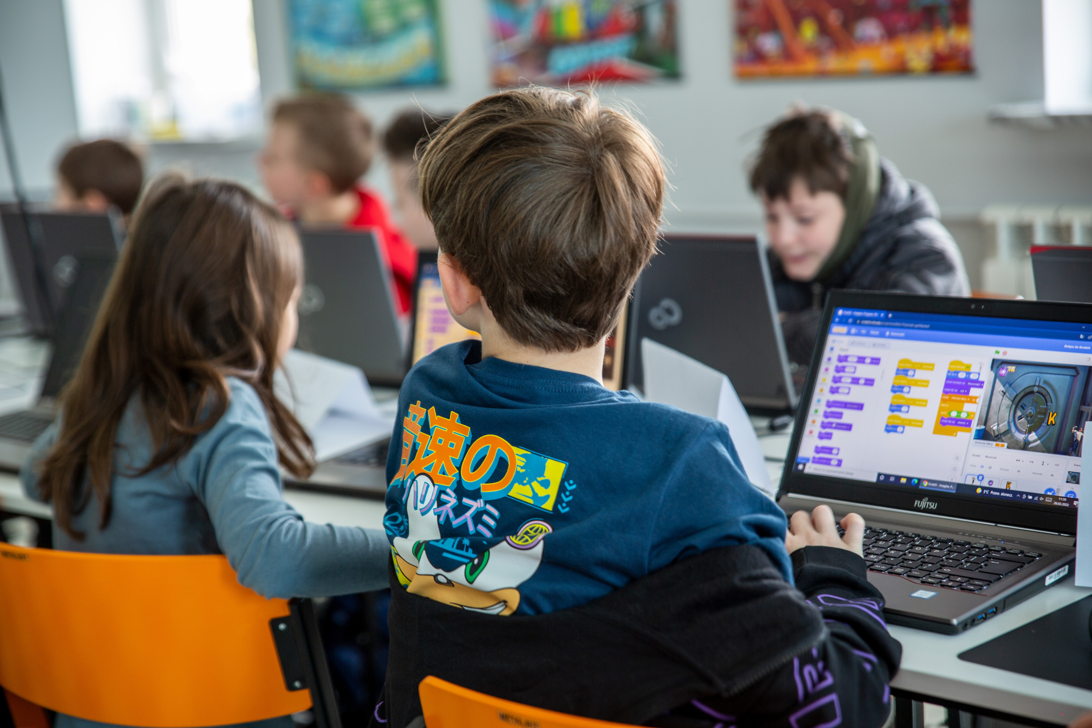 Dzieci siedzące przed komputerami w&nbsp;pracowni, na&nbsp;komputerach wyświetlają się kolorowe elementy
