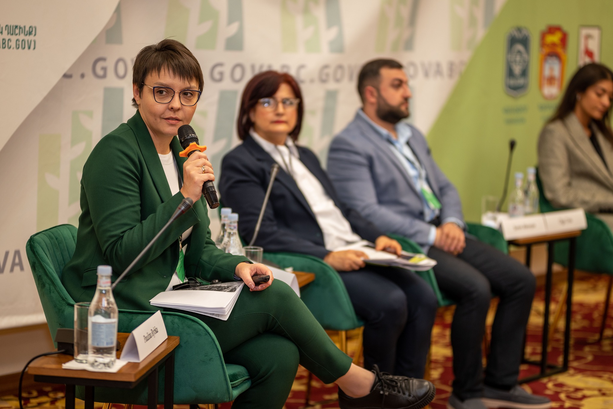 kobieta w zielonym garniturze siedzi na krześle, trzyma mikrofon, obok siedzą inne osoby