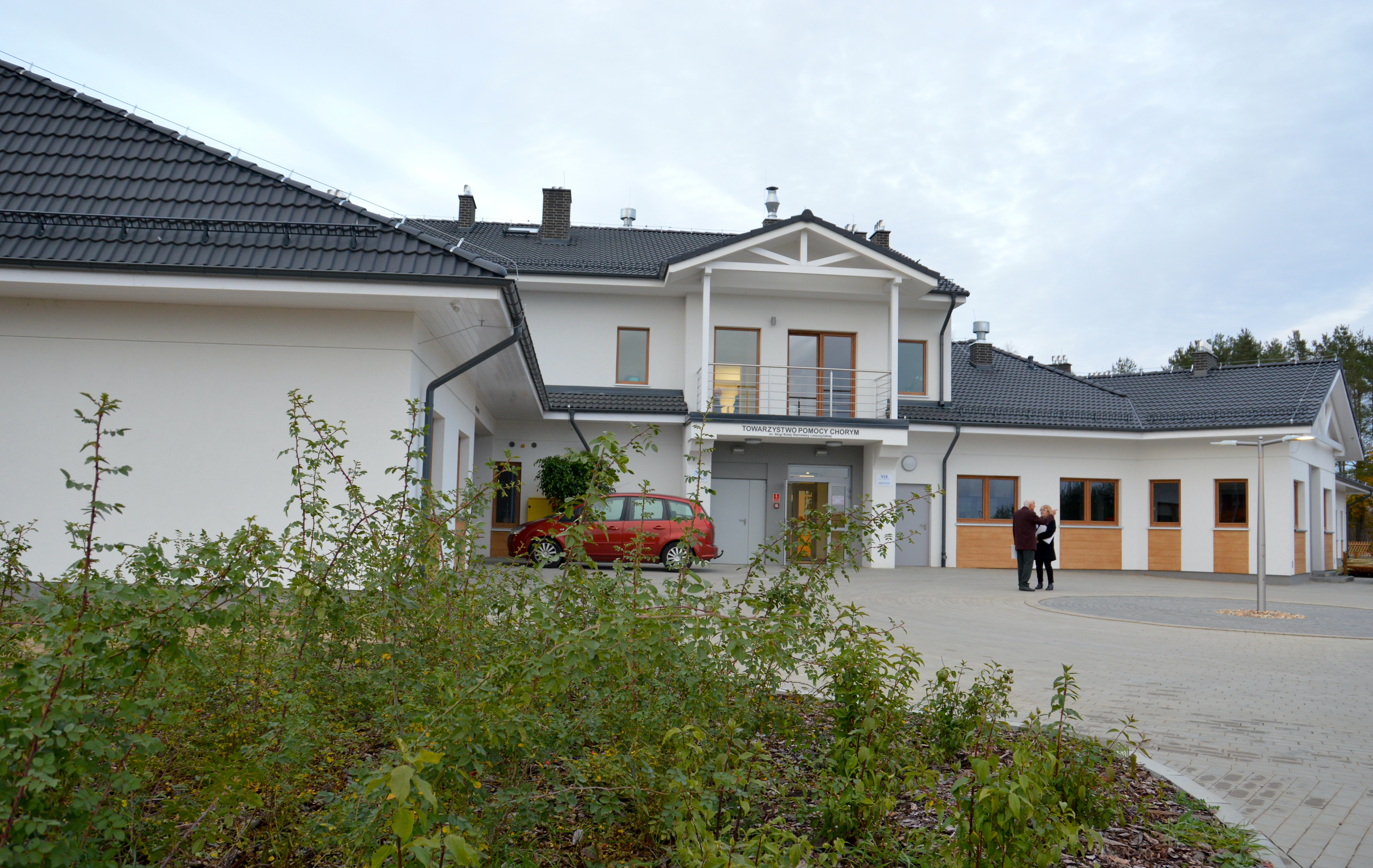 Parterowy biały budynek, przed nim dwoje rozmawiających ludzi i&nbsp;czerwone auto osobowe. Na pierwszym planie rabata roślin.