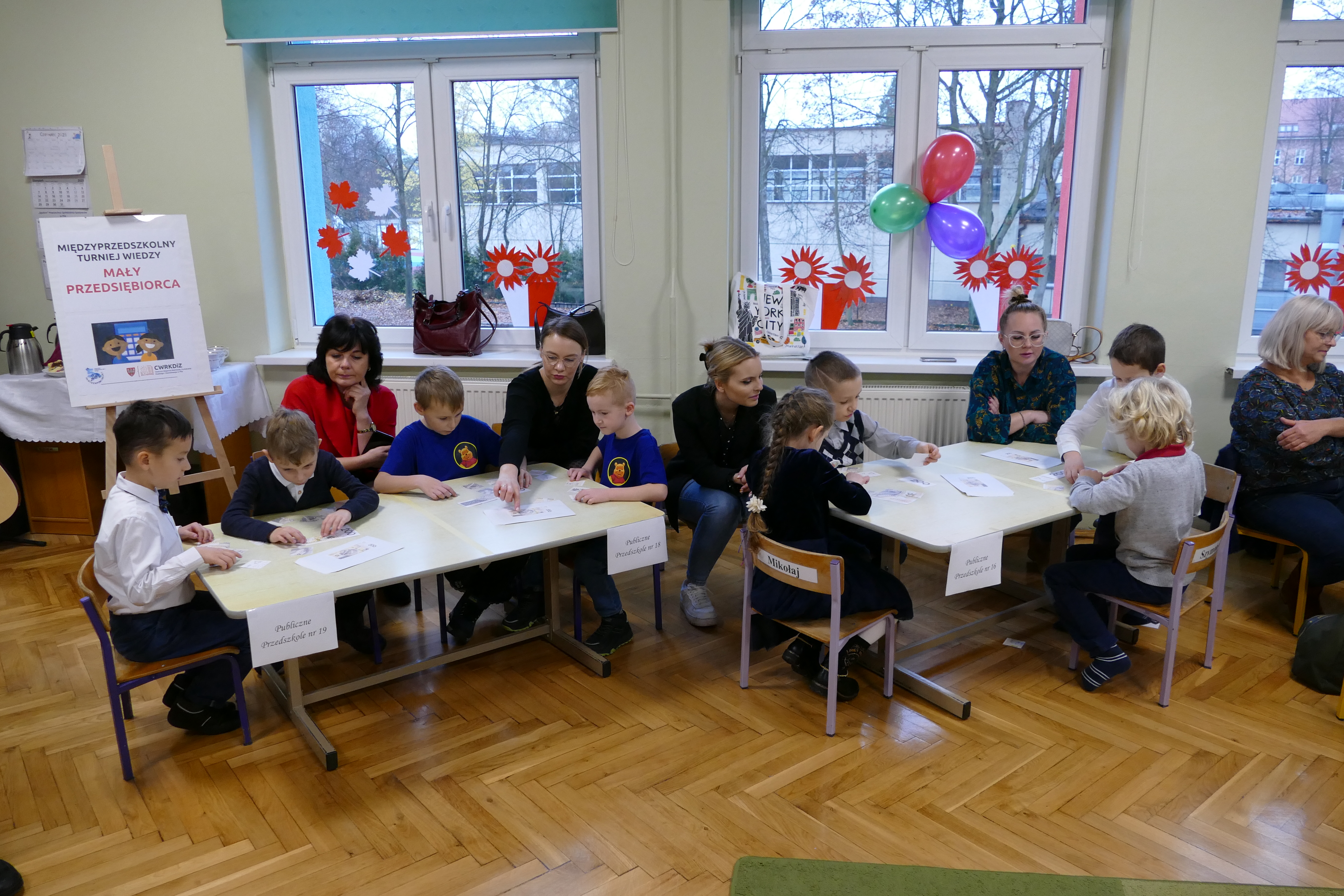 Sala przedszkolna, dzieci przy stolikach pracują nad zadaniami, na&nbsp;stolikach leżą karty pracy. W tle na&nbsp;sztaludze plakat z&nbsp;napisem "Międzyprzedszkolny turniej wiedzy mały przedsiębiorca"