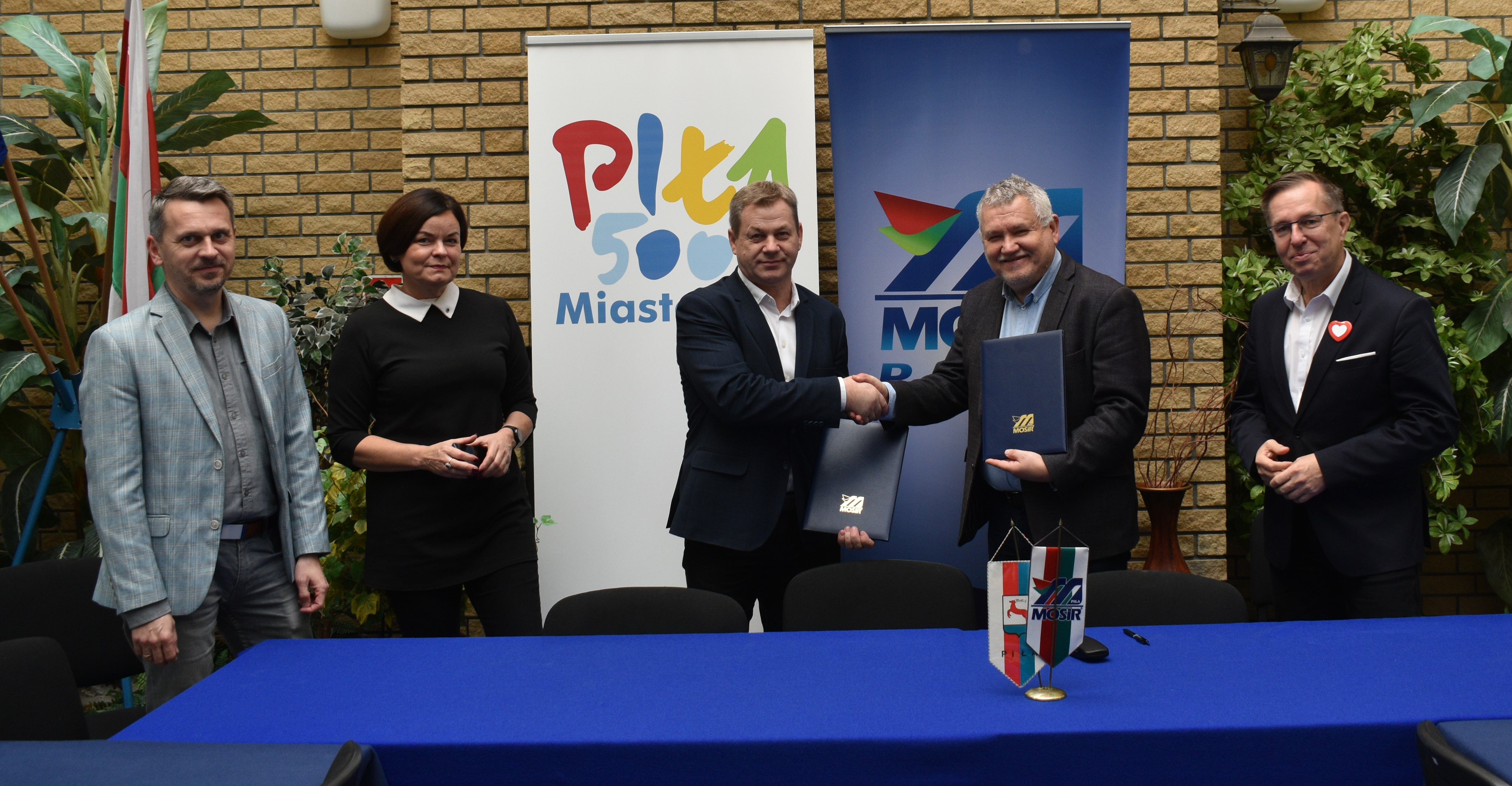 Pięc os&oacute;b: czterech mężczyn i&nbsp;jedna kobieta stoją w&nbsp;reprezentacyjnyn pomieszczeniu. Dw&oacute;ch mężczyn w&nbsp;środku ściska sobie dłonie i&nbsp;podaje teczki.