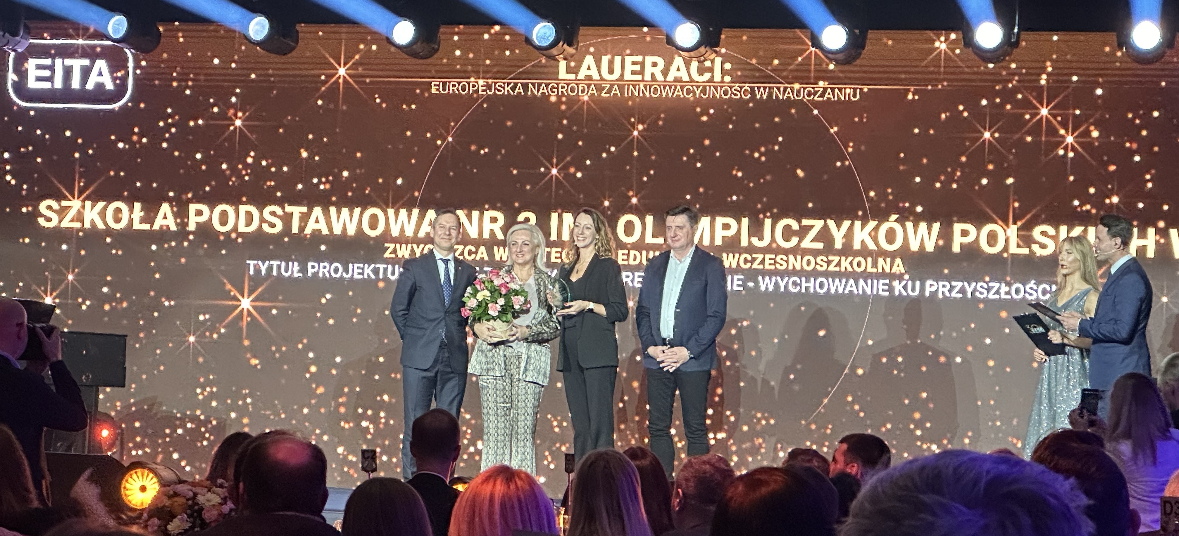  Dwie kobiety i&nbsp;dw&oacute;ch mężczyzn na&nbsp;scenie. Kobiety trzymają i&nbsp;prezentują statuetkę. W tle napis: Laureaci. Europejska nagroda za innowacyjność w&nbsp;nauczaniu. Szkoła Podstawowa nr 2 im. Olimpijczyk&oacute;w Polskich w&nbsp;Pile. Kategoria Edukacja wczesnoszkolna.
