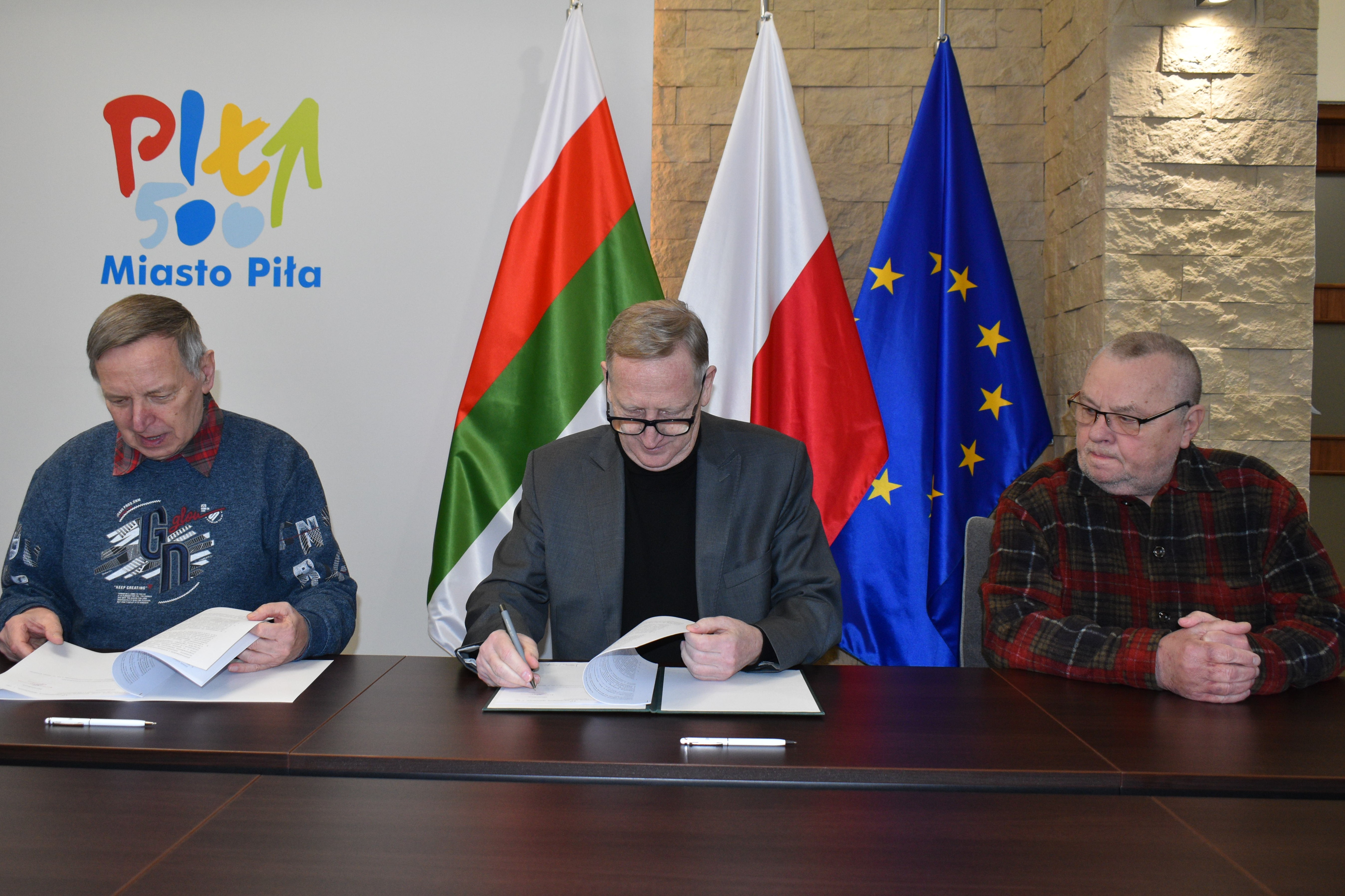 Soboty na dwóch kólkach 2023 Trzech mężczyn siedzi przy stole w reprezentacyjnym pomieszczeniu i podpisuje dokumenty. W tle logo Miasto Piła oraz falgi: Piły, Polski i Unii Europejskiej.