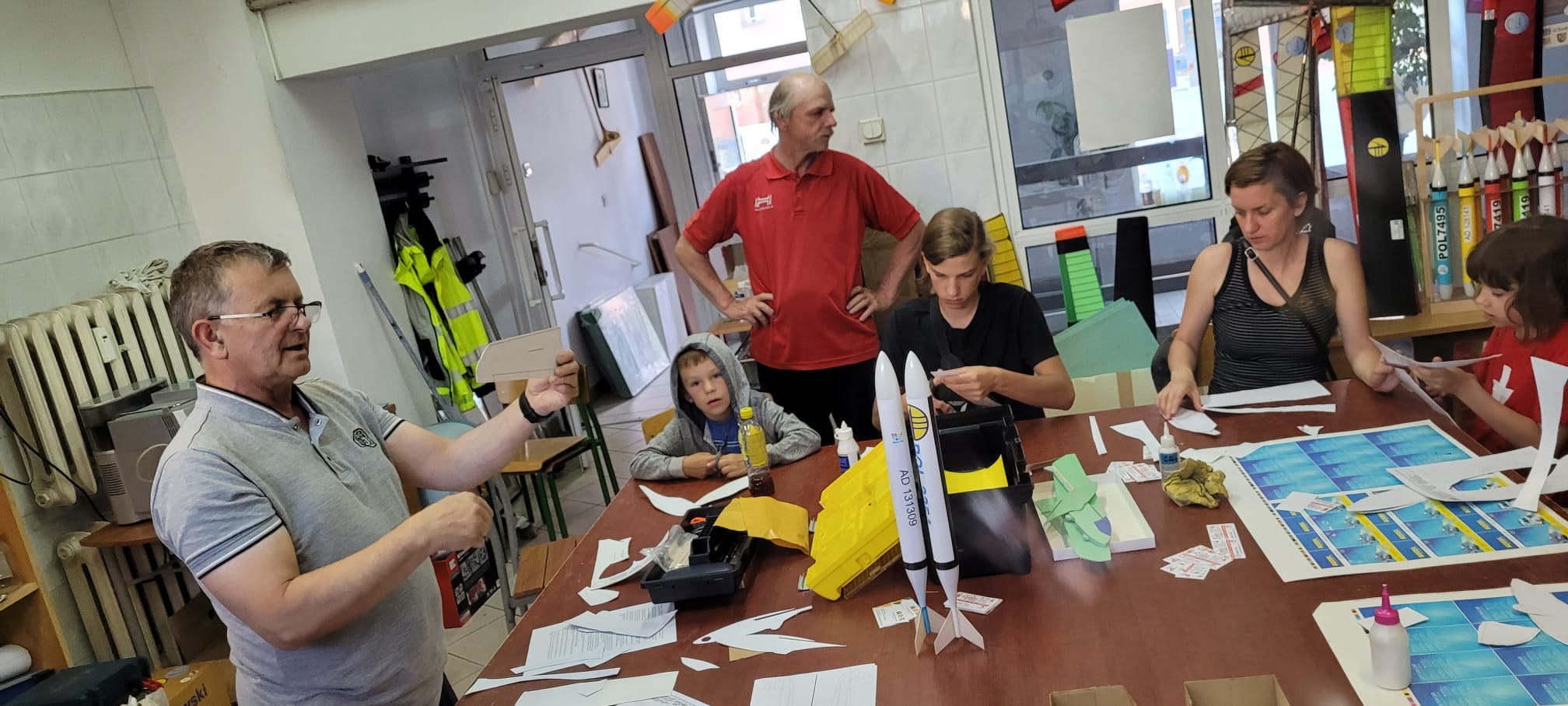 Pilski Klub Modelarzy Lotniczych Grupa dorosłych i dzieci w pracowni, na stole rakieta z papieru obok narzędzia, kleje i pomoce do robienia modeli latających.