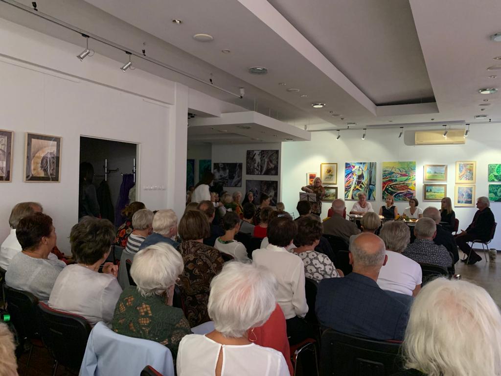 Grupa ludzi na wernisażu sztuki. Na ścianach wiszą obrazy, goście siedzą na krzesłach, w tle widać stoliki ze świecami, przy których siedzą artyści, obok kobieta gra na skrzypcach.