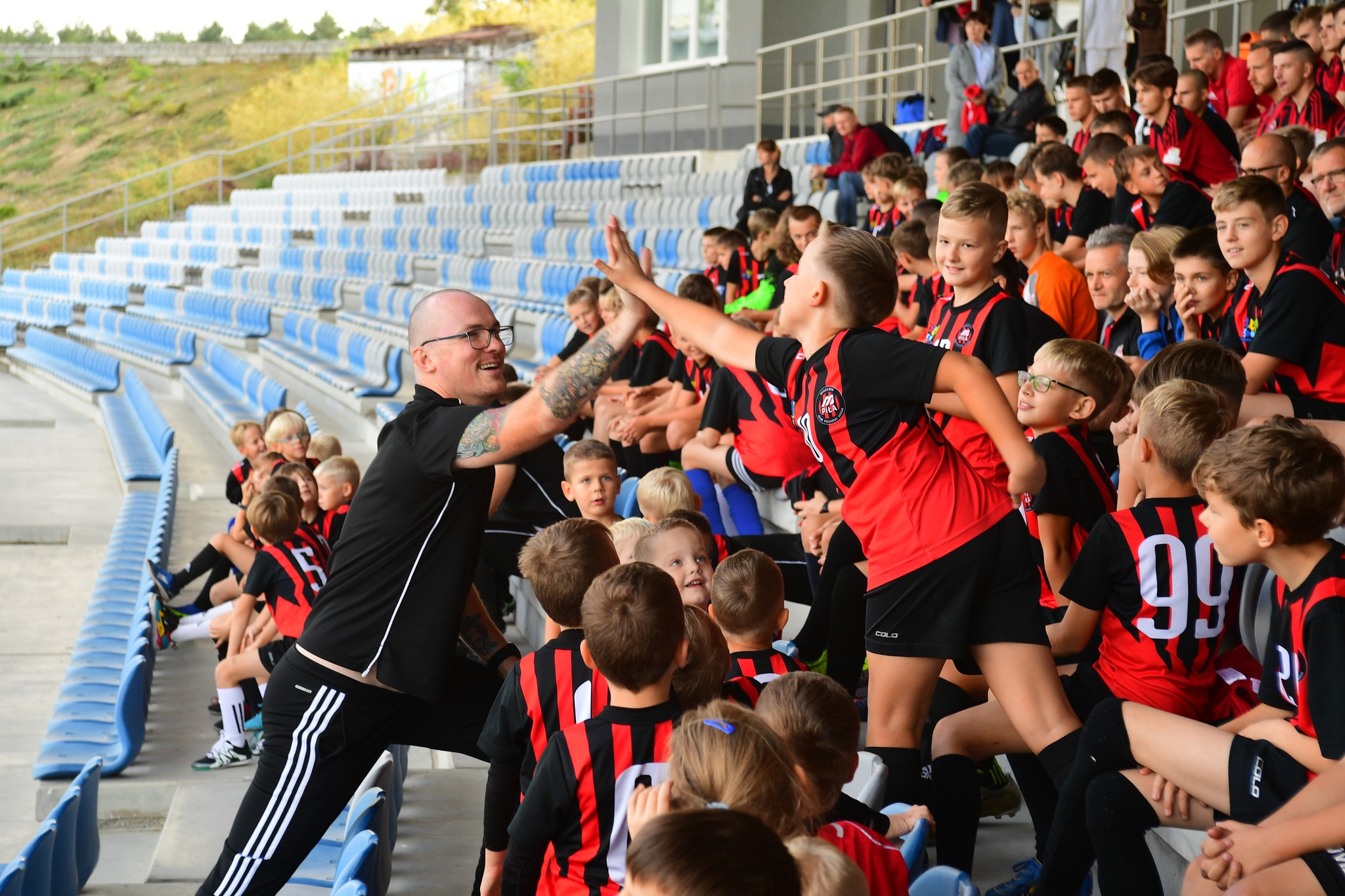Trybuny na stadionie, grupa chłopców w jednakowych czerwono-czarnych strojach sportowych, jeden z chłopców przybija piątkę z trenerem.