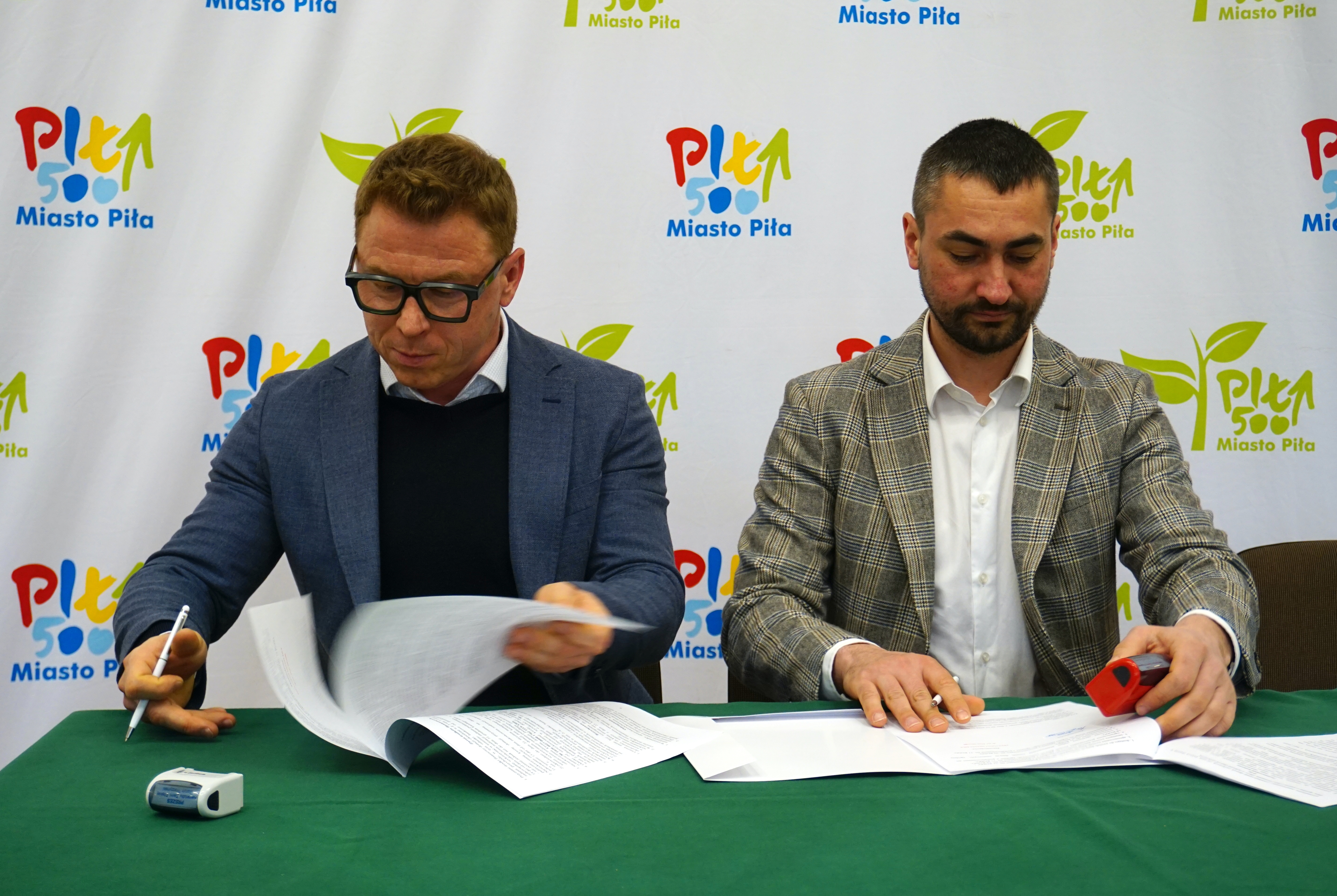 Dwóch mężczyzn przy stole podpisuje dokumenty, za nimi na ściance logotypy Miasto Piła.
