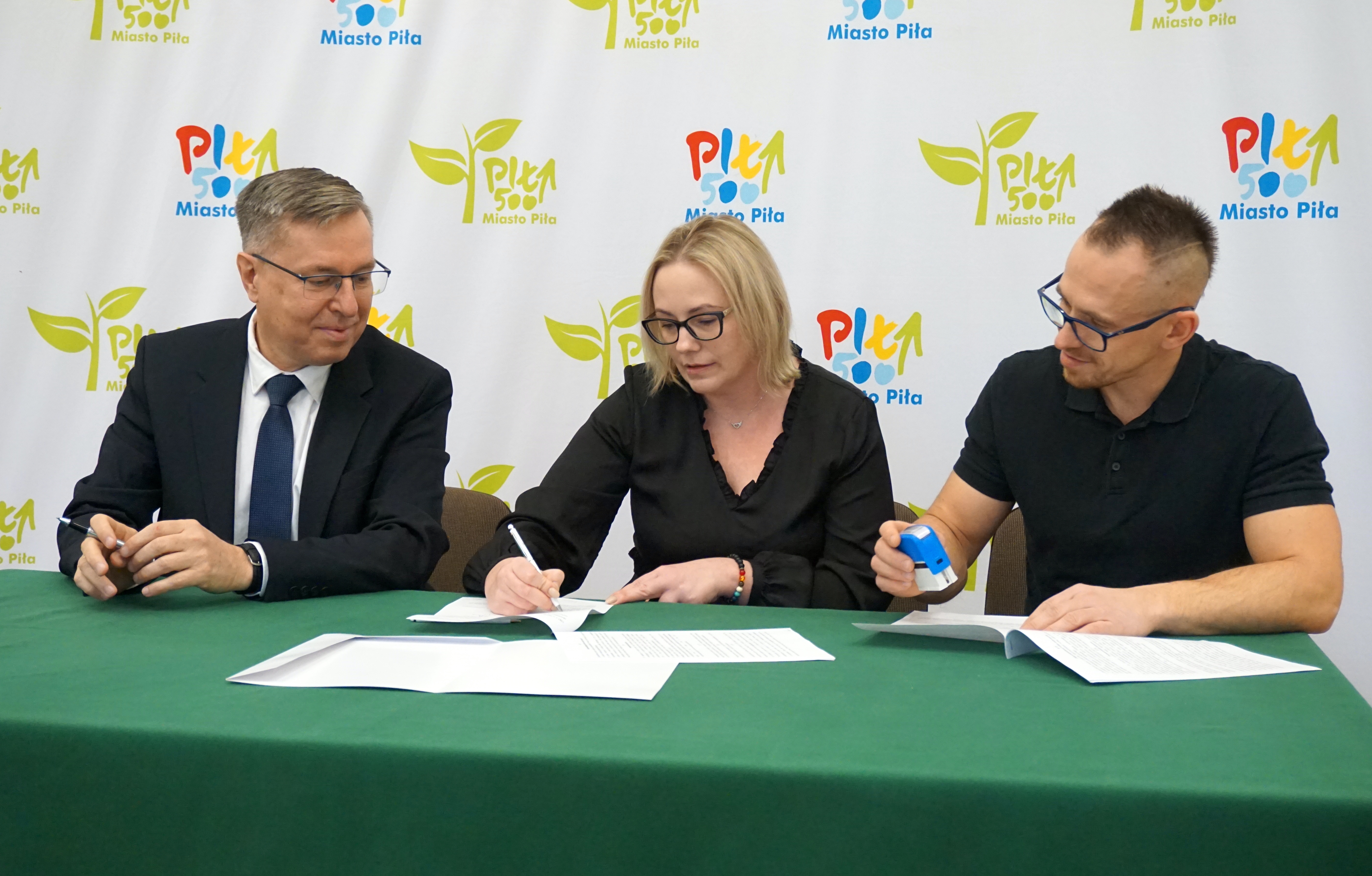 Kobieta i dwóch mężczyzn podpisują dokumenty siedząc przy stole, w tle ścianka z logotypami Miasto Piła.