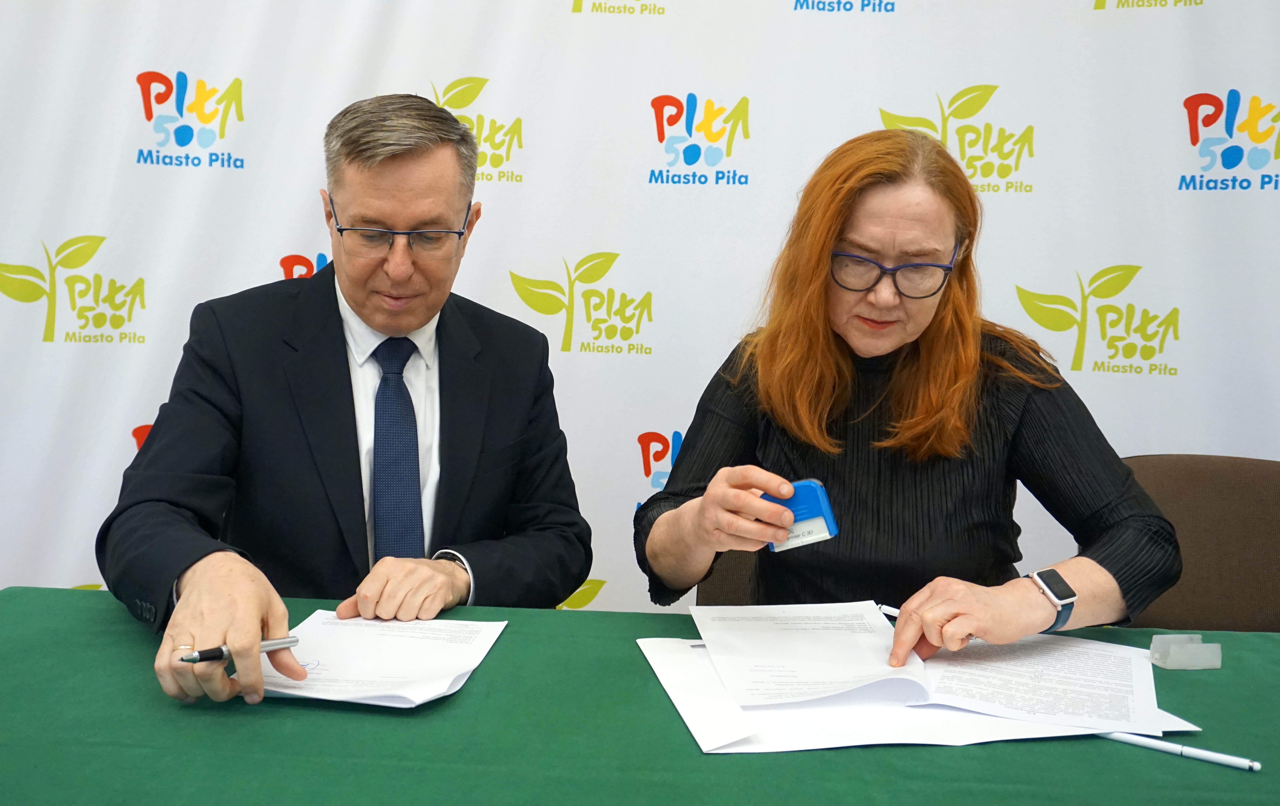 Kobieta i mężczyzna siedzą przy stole w pomieszczeniu, podpisują dokumenty. W tle ścianka z logotypami Miasto Piła.