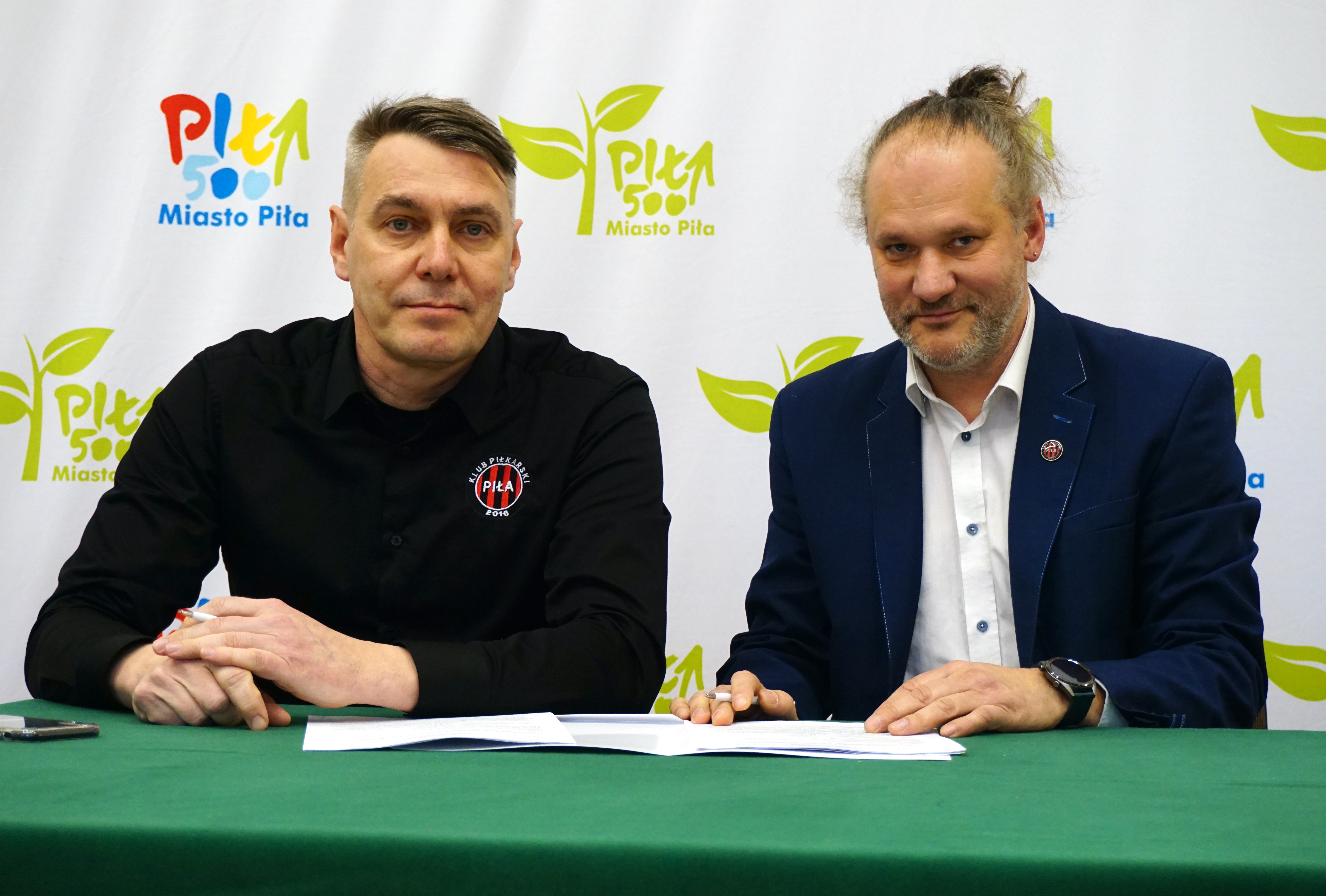 Dwóch mężczyzn przy stole podpisuje dokumenty, w tle ścianka z logotypami miasto piła.