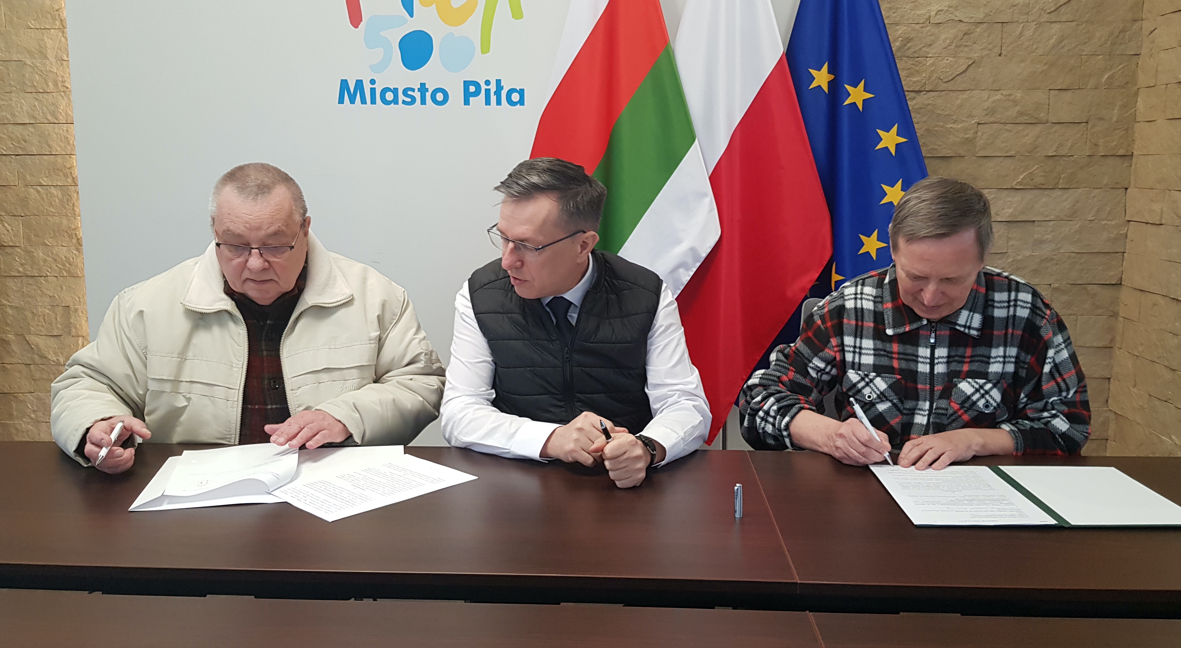 Zapraszamy na 18 rajd Wiosenne Przebudzenie! Trzech mężczyzn siedzi przy stole, podpisują dokumenty. Na ścianie logotyp Miasto Piła, flaga Piły, Polski i UE.