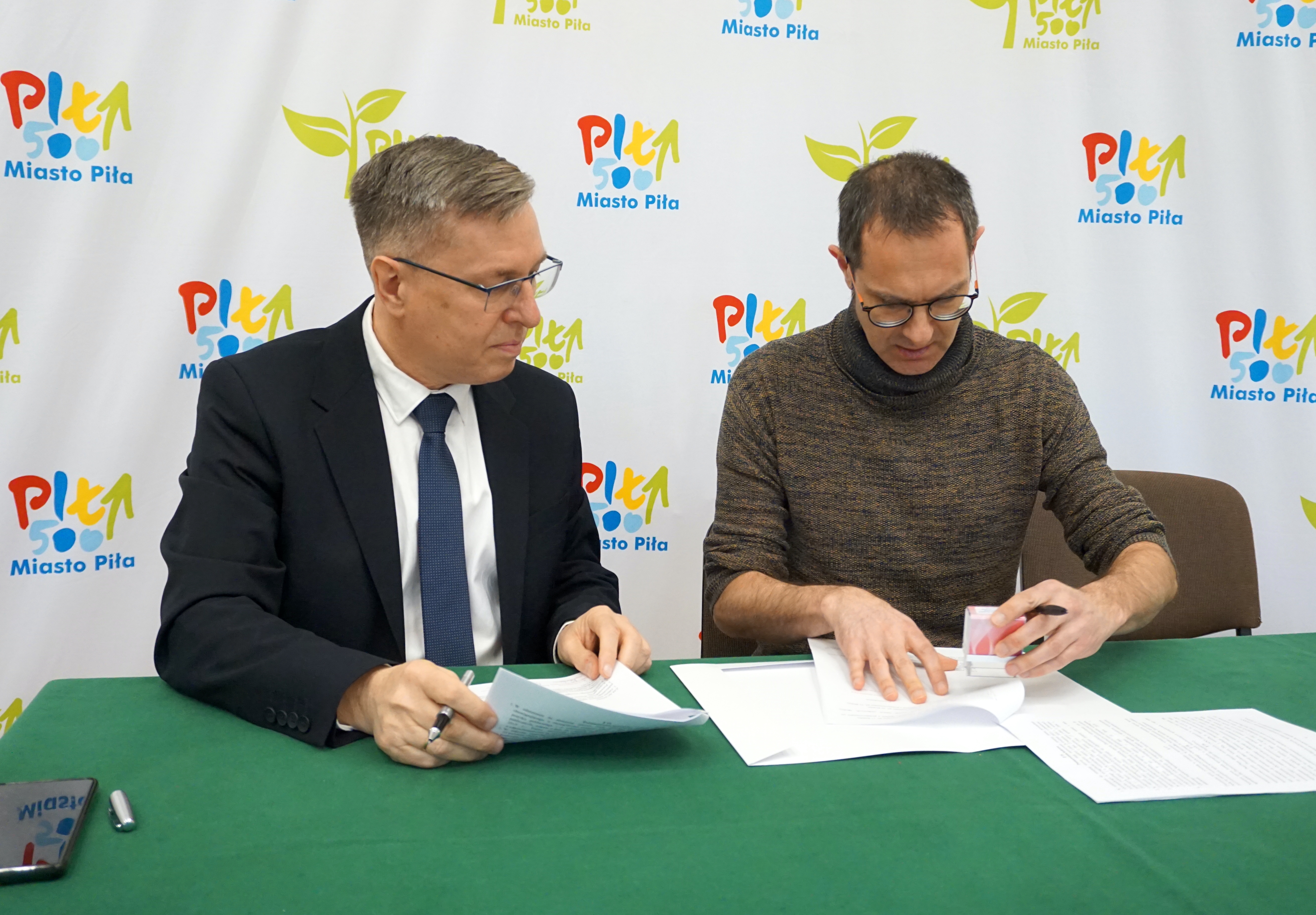 Dwóch mężczyzn w&nbsp;pomieszczeniu podpisuje dokumenty siedząc przy stole, w&nbsp;tle ścianka z&nbsp;logotypami miasto Piła.