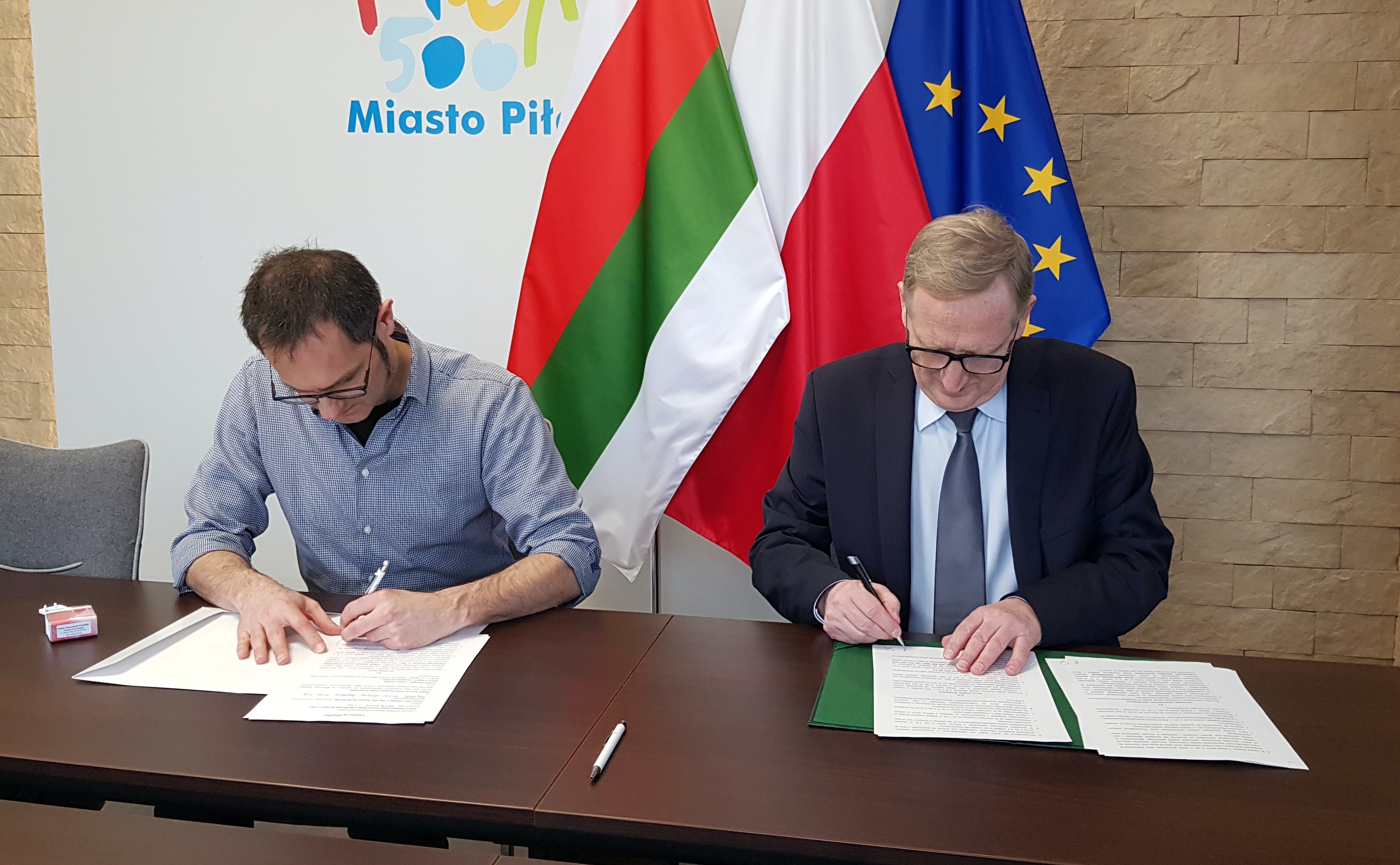 Wizyta miasta partnerskiego Châtellerault Dwóch mężczyzn siedzi przy stole, podpisują dokumenty. W tle flaga Piły, Polski i Unii Europejskiej. Na ścianie logotyp Miasto Piła.