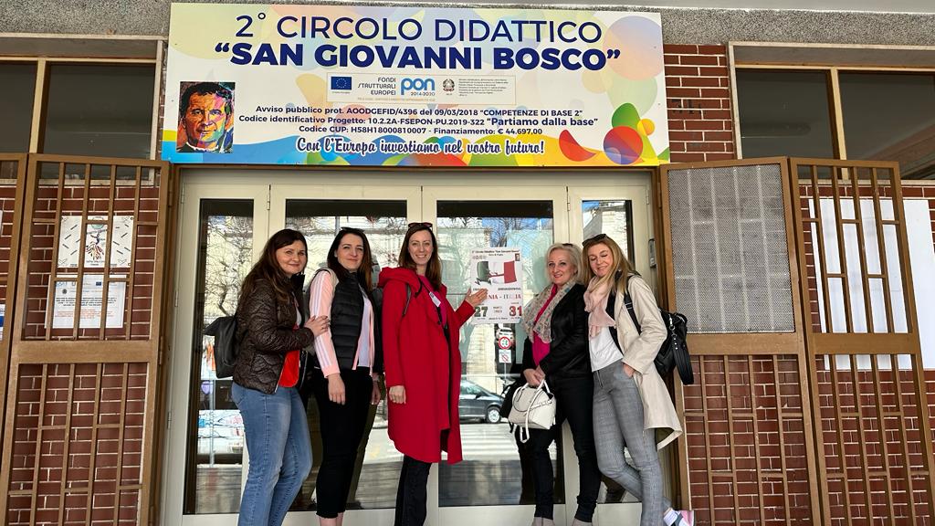 Pięć kobiet stoi przed drzwiami wejściowymi do budynku. Nad drzwiami widnieje napis w&nbsp;języku włoskim: "2 CIRCOLO DIDATTTICO SAN GIOVANNI DOSCO