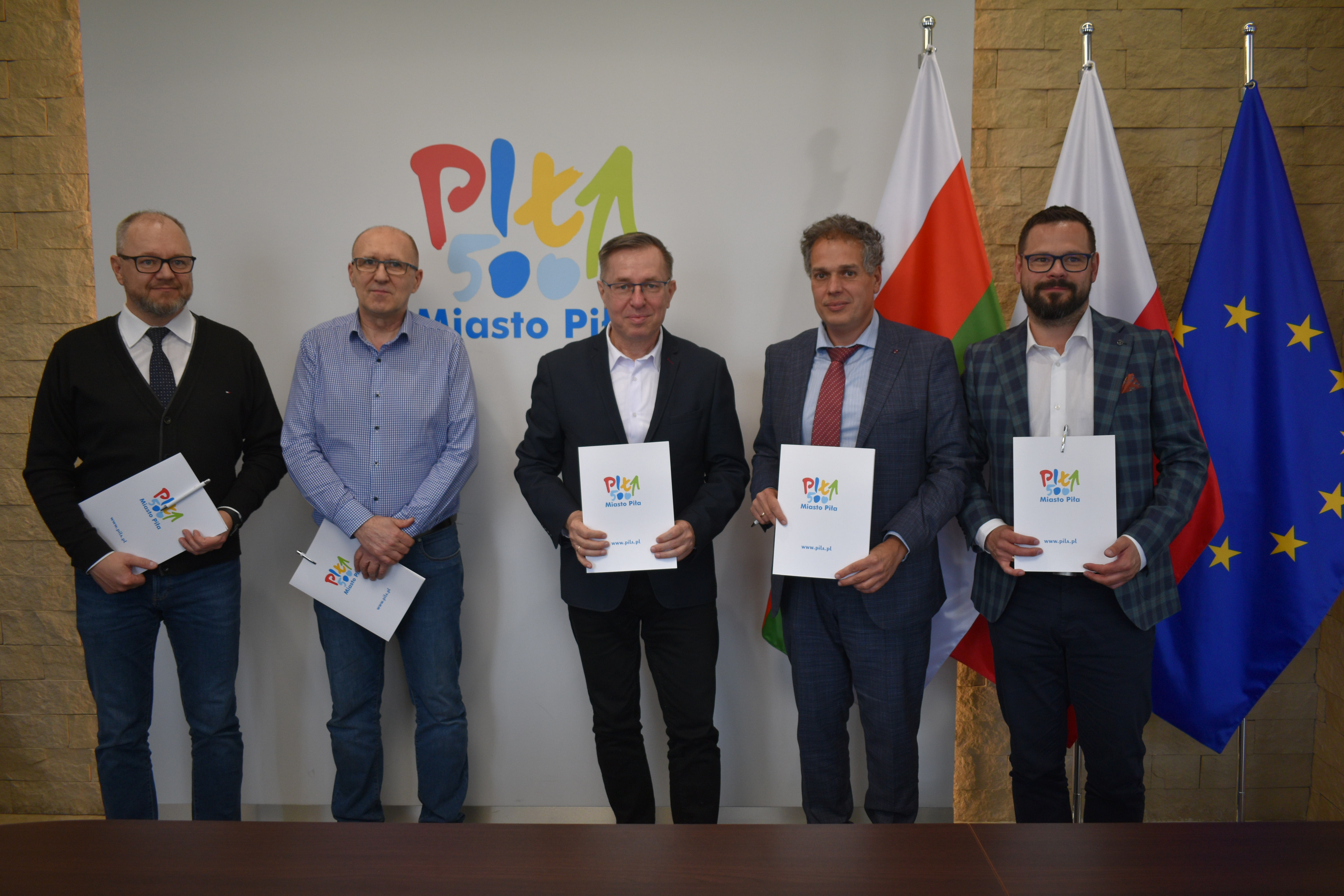 Pięciu mężczyn stoi w&nbsp;reprezentacyjnym pomieszczeniu. W dłoniach trzymają białe teczki z&nbsp;logotypem Miasta Piły. W tle flagi: Polska, Piły i&nbsp;Unii Europejskiej.