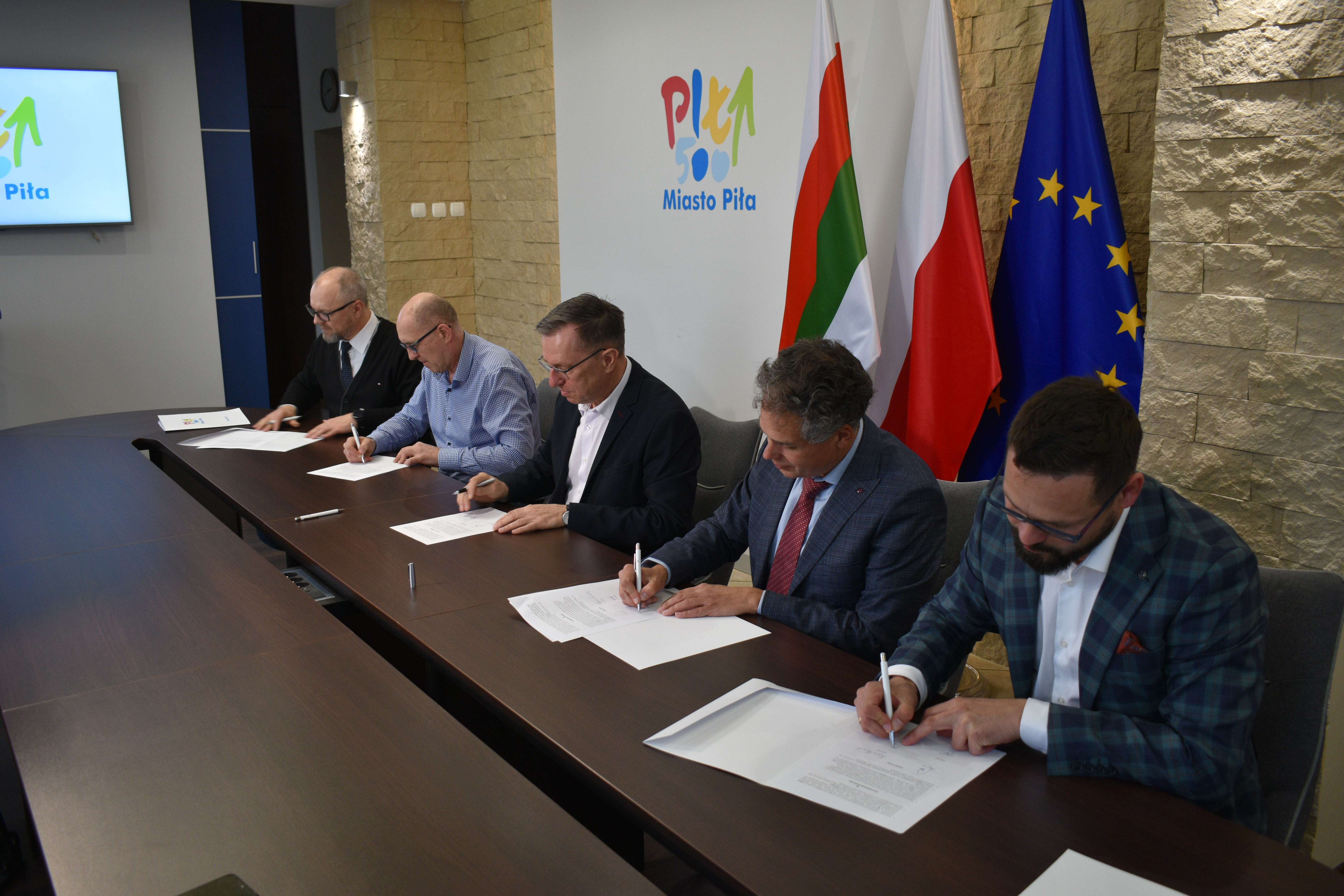 Pięciu mężczzn siedzi przy stole w&nbsp;reprezentacyjnym pomieszczeniu i&nbsp;podpisuje dokumenty. W tle flagi: Polska, Piły i&nbsp;Unii Europejskiej.