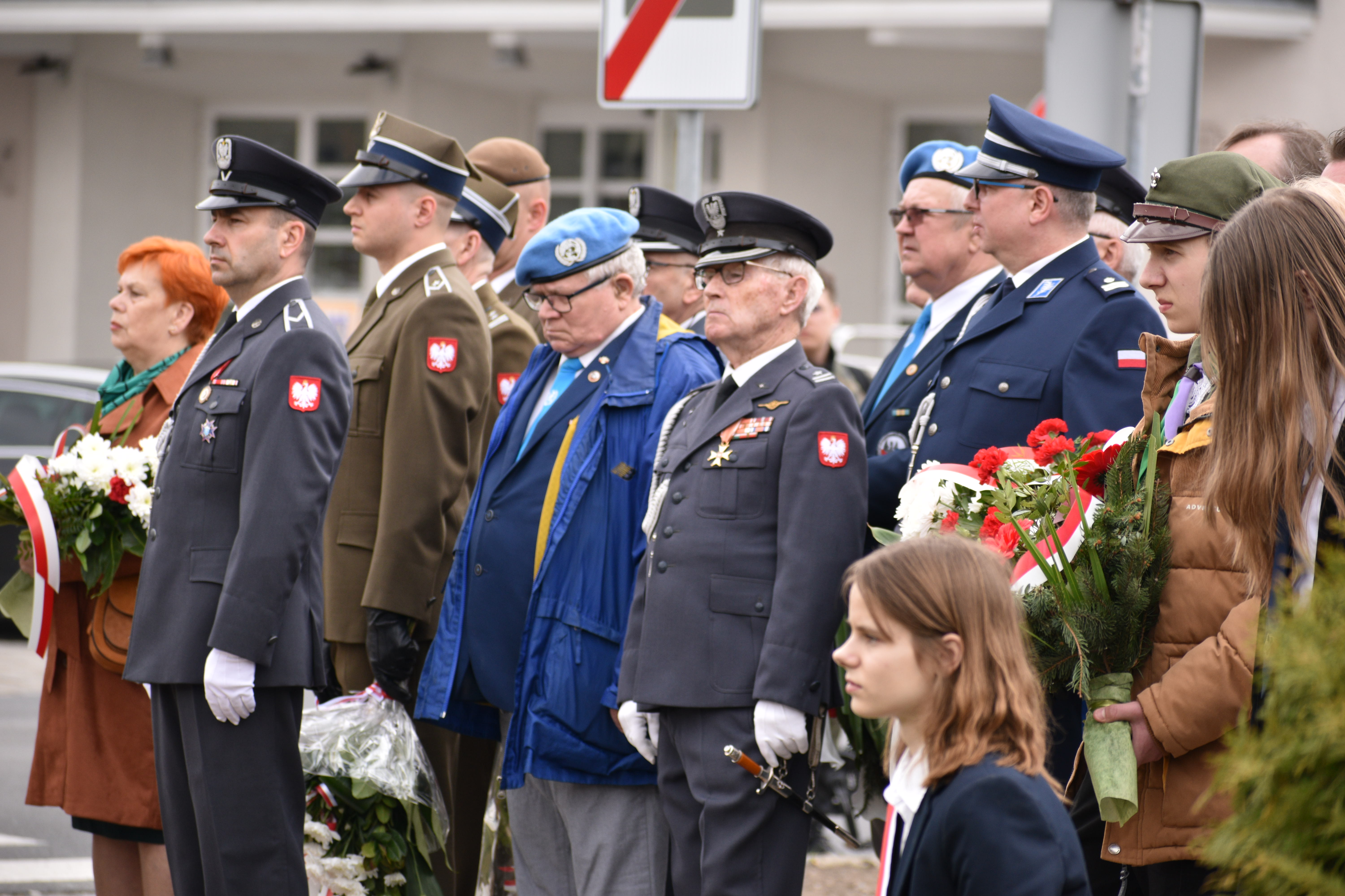 Grupa ludzi, mundurowych stoi na&nbsp;świeżym powietrzu. Kilka osób trzyma wiązanki biało-czerwone.