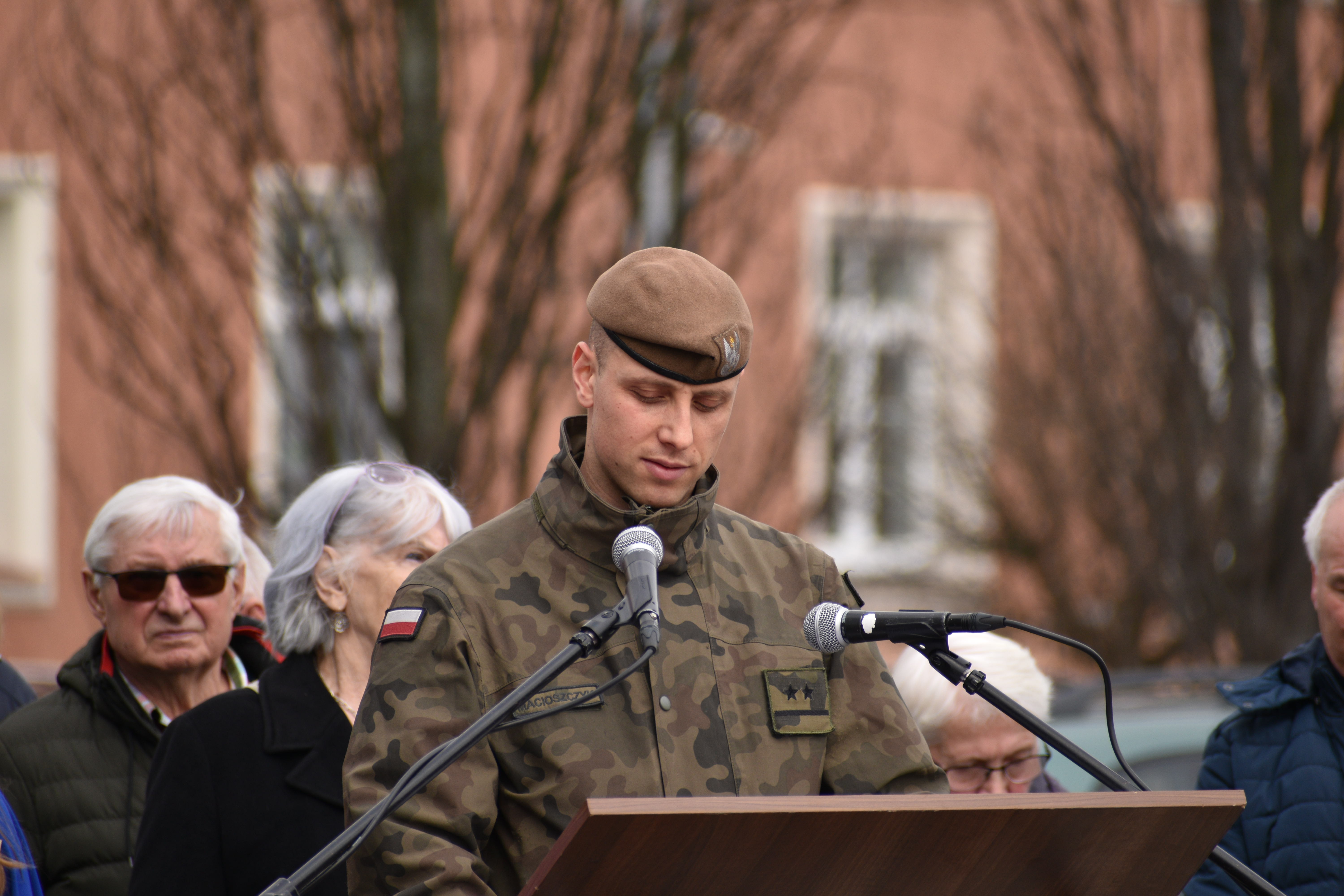 Żołnierz przy mównicy, spogląda w&nbsp;dół. W tle ludzie.