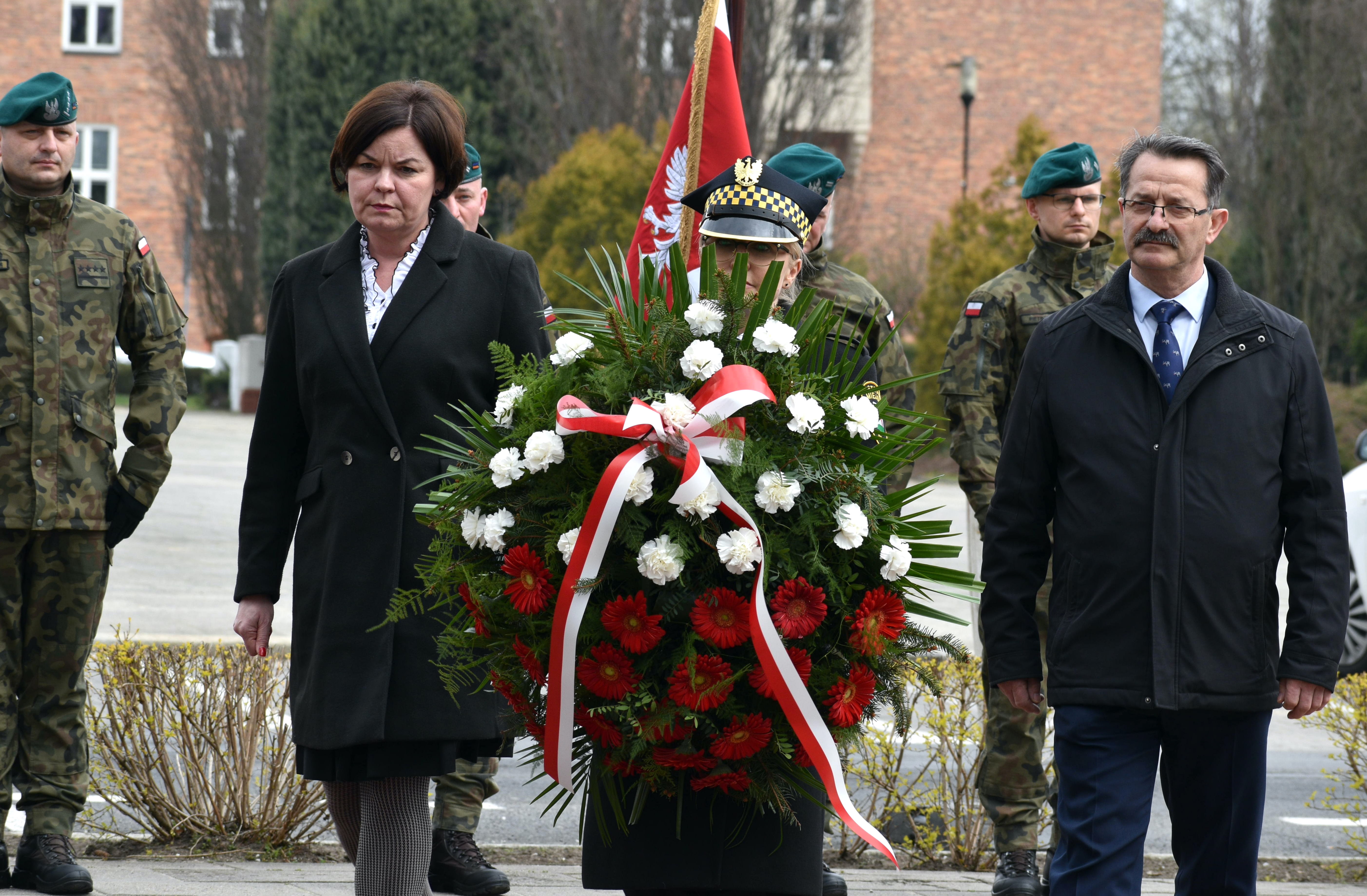 Kobieta, mężczyzna i&nbsp;strażnik miejski niosą wiązankę kwiatów w&nbsp;biało-czerwonych barwach, w&nbsp;tle żołnierze i&nbsp;budynek.