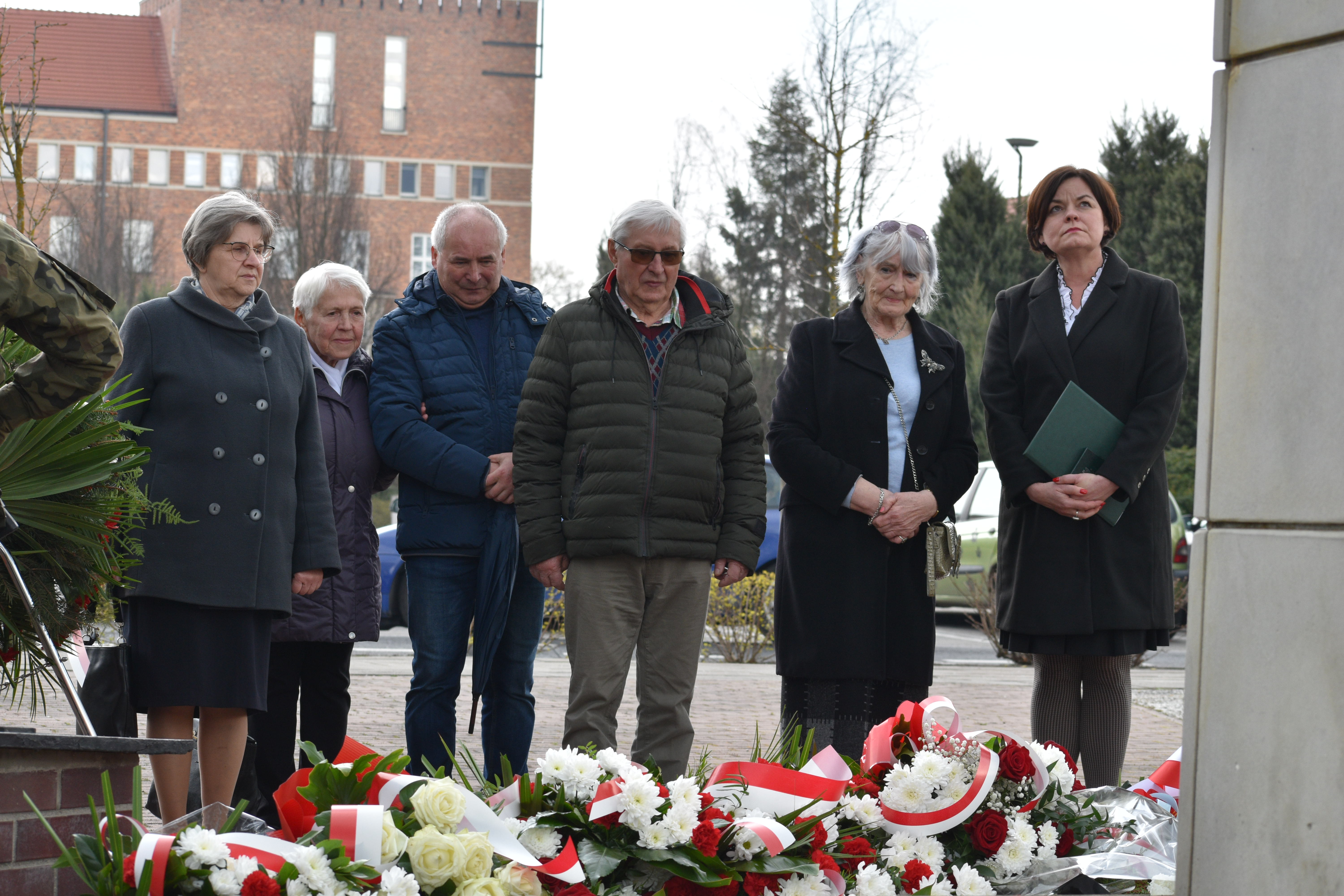 Grupa osób stoi przed pomnikiem. Widać złożone wiązanki kwiatów. W tle budynek.