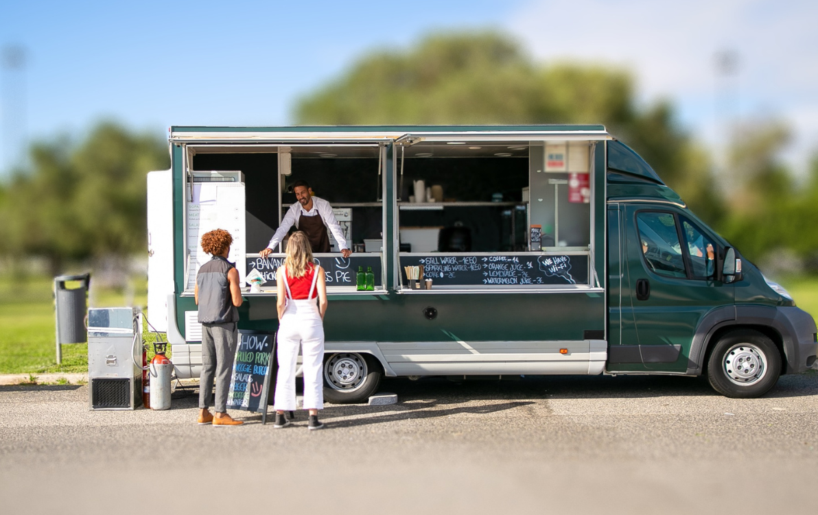Postaw food truck na Bulwarach Europejskich! Foodtruck stojący w przestrzeni miasta, przed autem stoją dwie osoby, reklama na potykaczu.