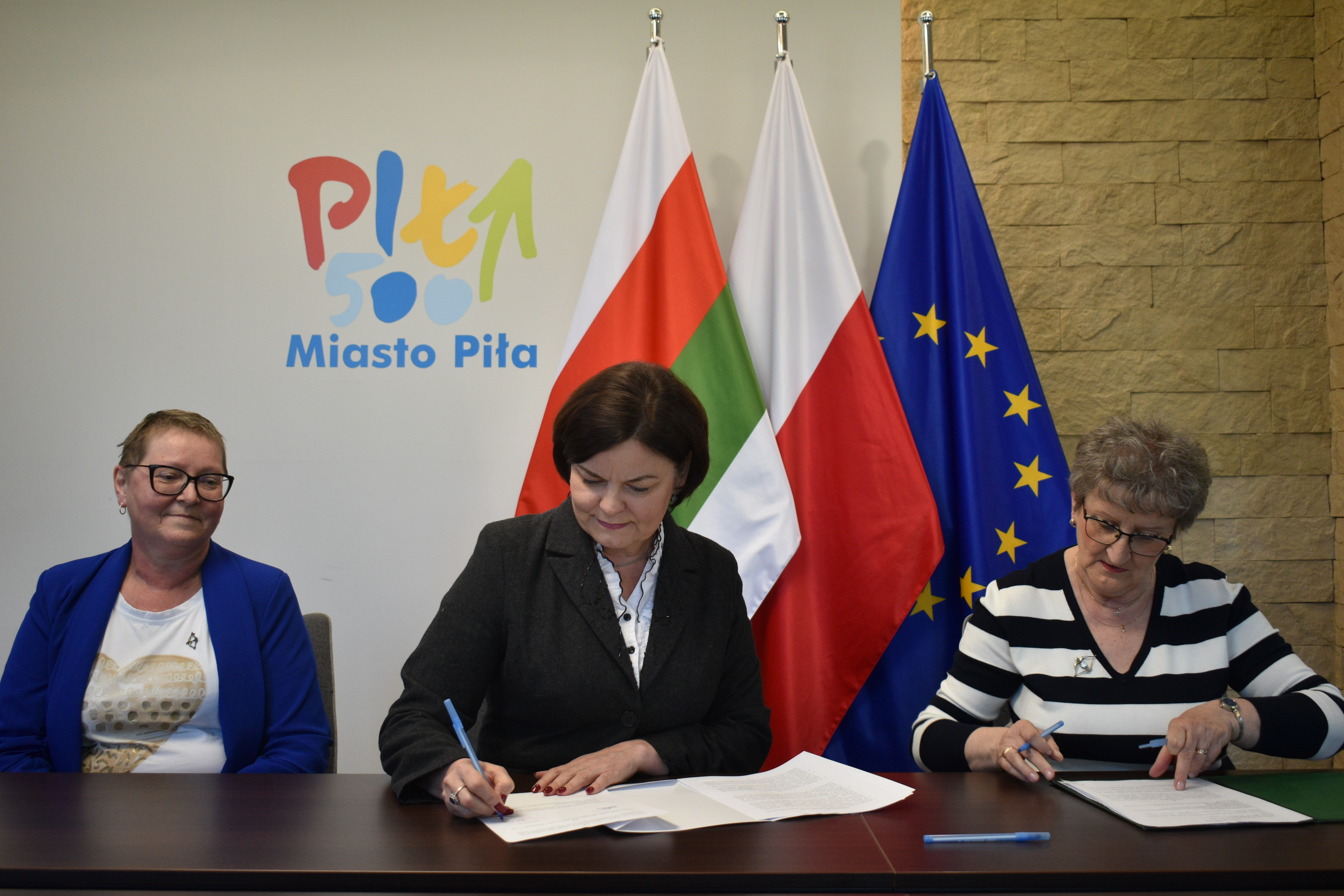 Trzy kobiety siedzą przy stole w&nbsp;reprezentacyjnym pomieszczeniu i&nbsp;podpisują dokumenty. W tle logotyp Miasto Piła i&nbsp;trzy flagi: Polski, Miasta Piły i&nbsp;Unii Europejskiej.