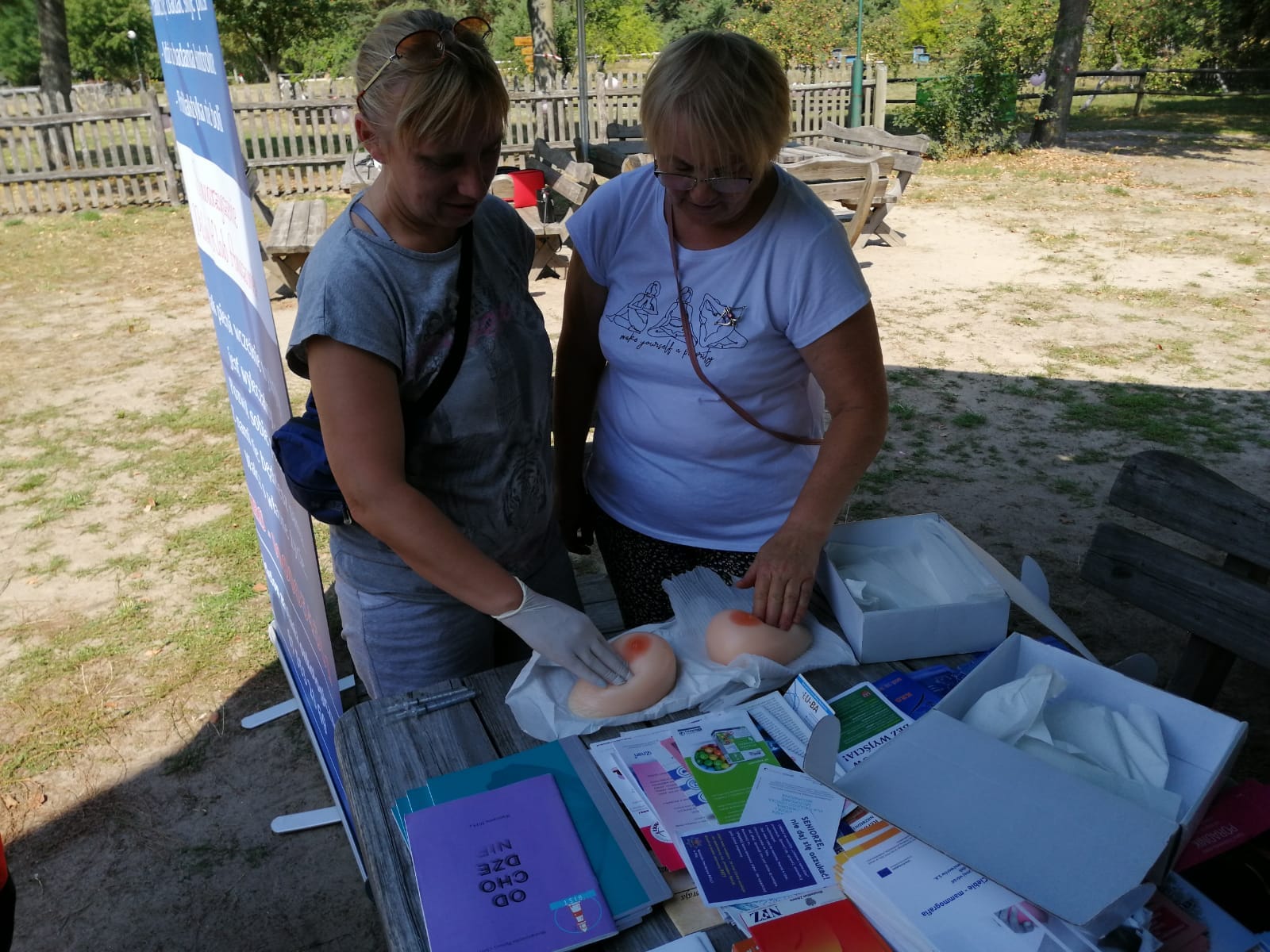 Dwie kobiety stoją przy stoliku ustawionym pod namiotem. Na stole ułożone fantomy służące do ćwiczenia samobadania piersi przez kobiety. Stojące przy stoliku kobiety ćwiczą. 