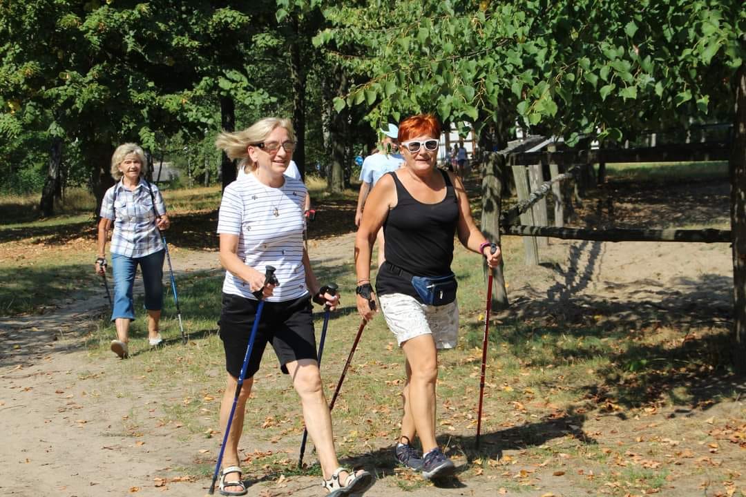 Trzy uśmiechnięte kobiety maszerują na&nbsp;świeżym powietrzu z&nbsp;kijkami Nordic Walking. W tle zieleń, drzewa.