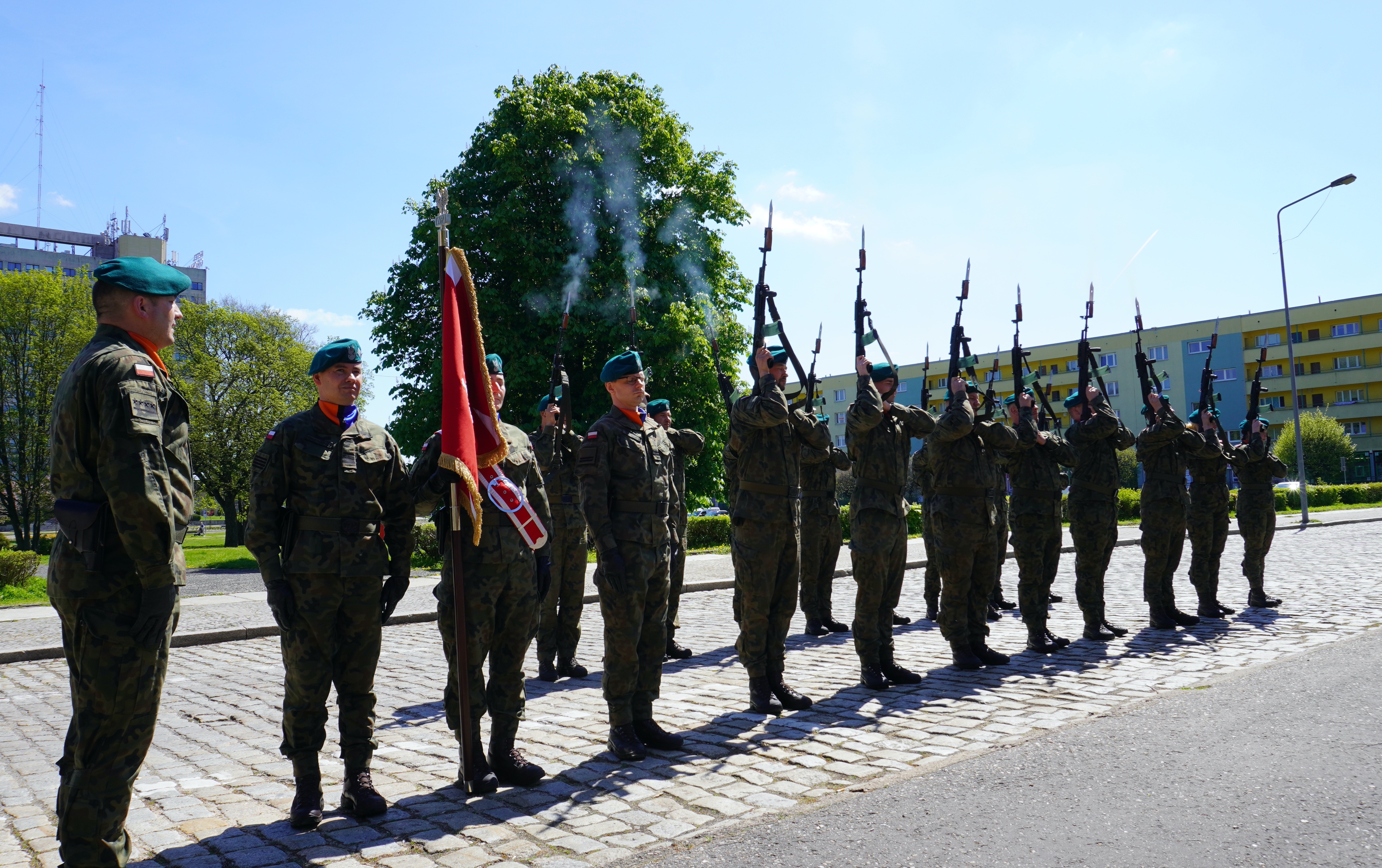 Grupa żołnierzy, jeden z nich trzyma sztandar, reszta oddaje strzał w ramach salwy honorowej. Przestrzeń miejska, zieleń, w tle zabudowania.