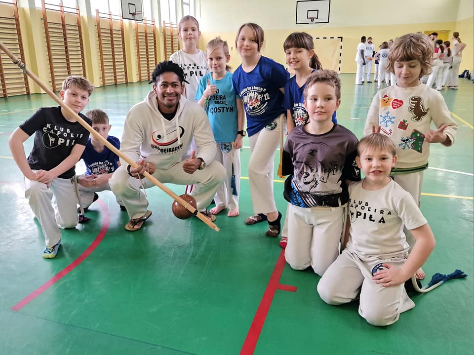 Grupa dzieci, w której są dziewczynki i chłopcy oraz jeden mężczyzna przebywają w sali gimnastycznej. Wszyscy mają ubrane stroje sportowe. Mężczyzna trzyma w dłoniach kij.