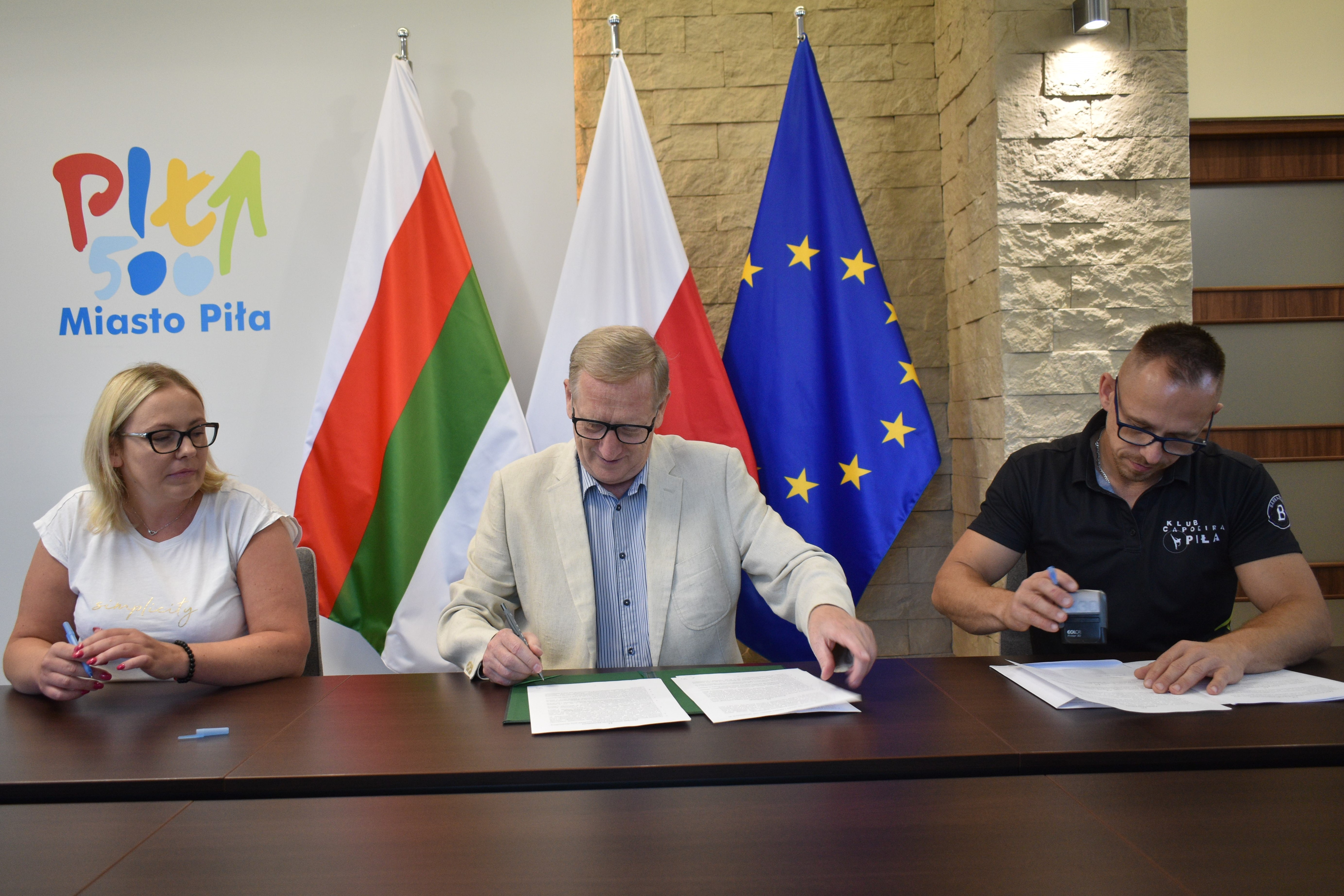 Kobieta i dwóch mężczyn siedzą za stołem w reprezentacyjnym pomieszczeniu i podpisują dokumenty. W tle logotyp Miast Piło i flagi: Polski, Piły i Unii Europejskiej.