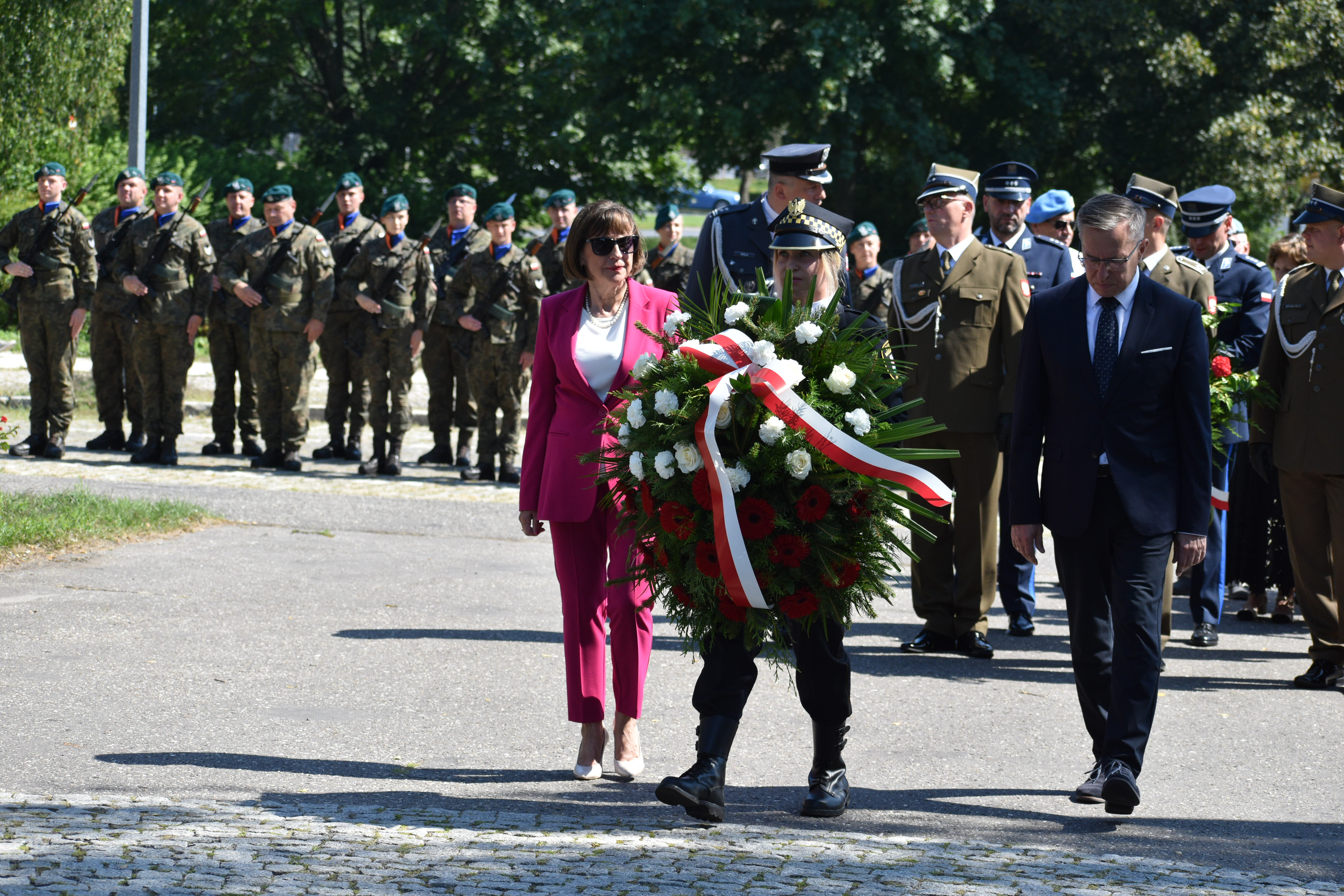 kobieta i&nbsp;mężczyzna idą za strażniczką miejską, która niesie wieniec kwiatów, w&nbsp;tle wojsko i&nbsp;inne delegacje/