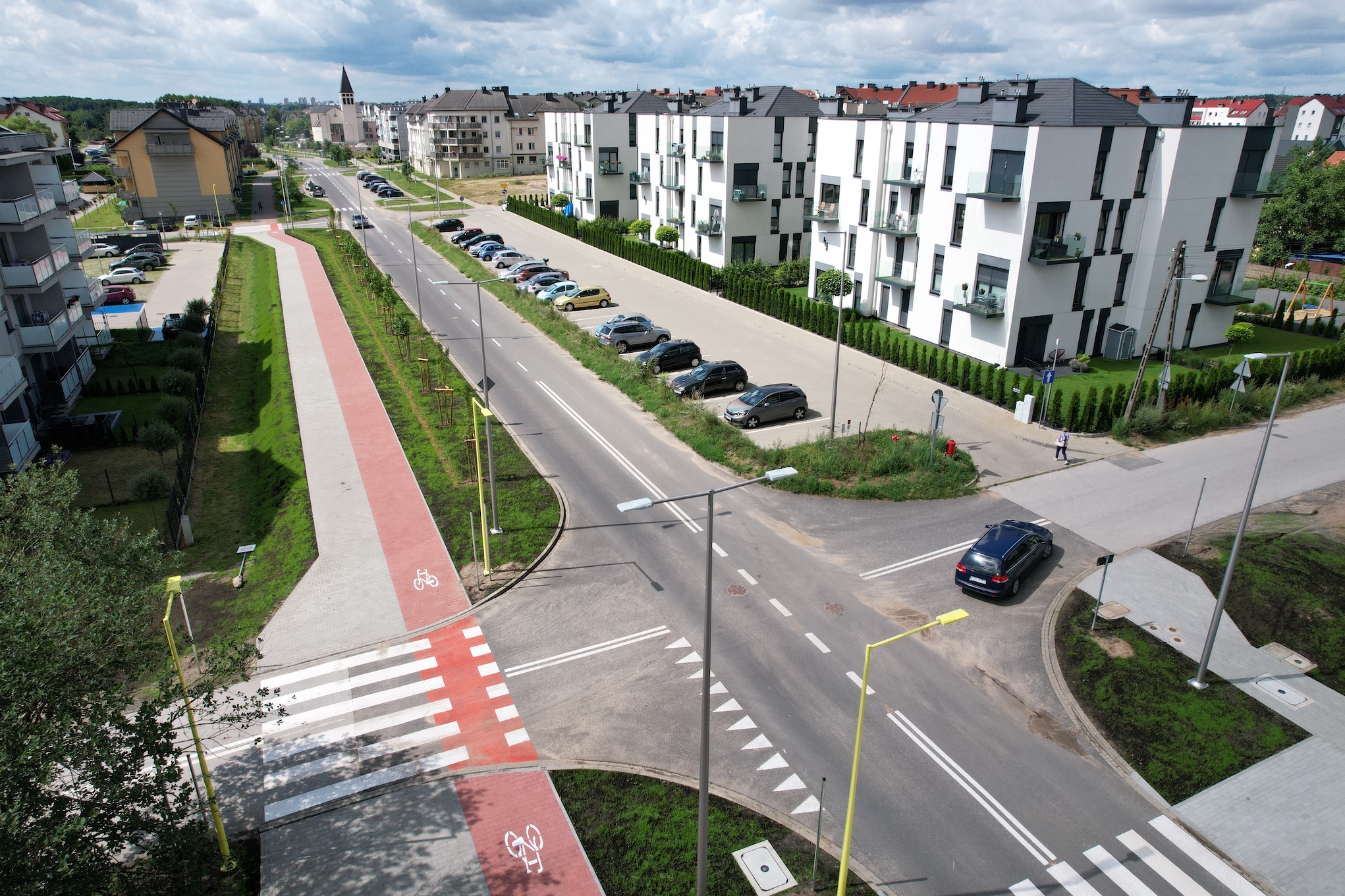 Zmiany na ulicy Kazimierza Wielkiego Zdjęcie z lotu ptaka. Miasto, środkiem przebiega główna ulica, obok chodnik i ścieżka rowerowa, w tle kościół. Po dwóch stronach zabudowania, bloki wielorodzinne, duży parking i auta osobowe, dużo zieleni.