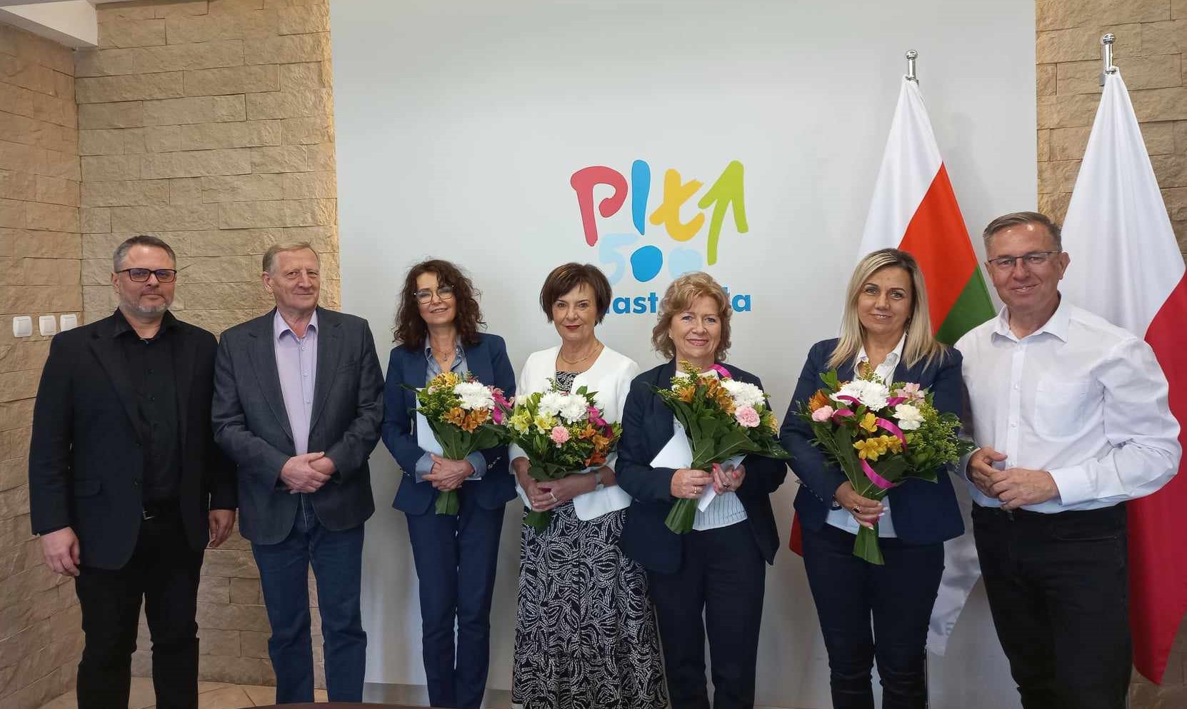 zdjęcie 4 kobiety i 3 mężczyzn pozują do zdjęcia na tle ściany z banerem, kobiety trzymają kwiaty.