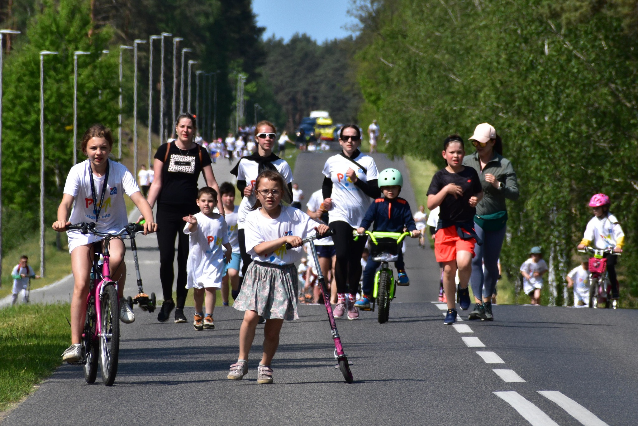 Droga wiodąca przez las, na której odbywają się zawody sportowe. Ludzie w róznym wieku: dzieci i dorośli biegną, maszerują, jadą rowerami i hulajnogami.