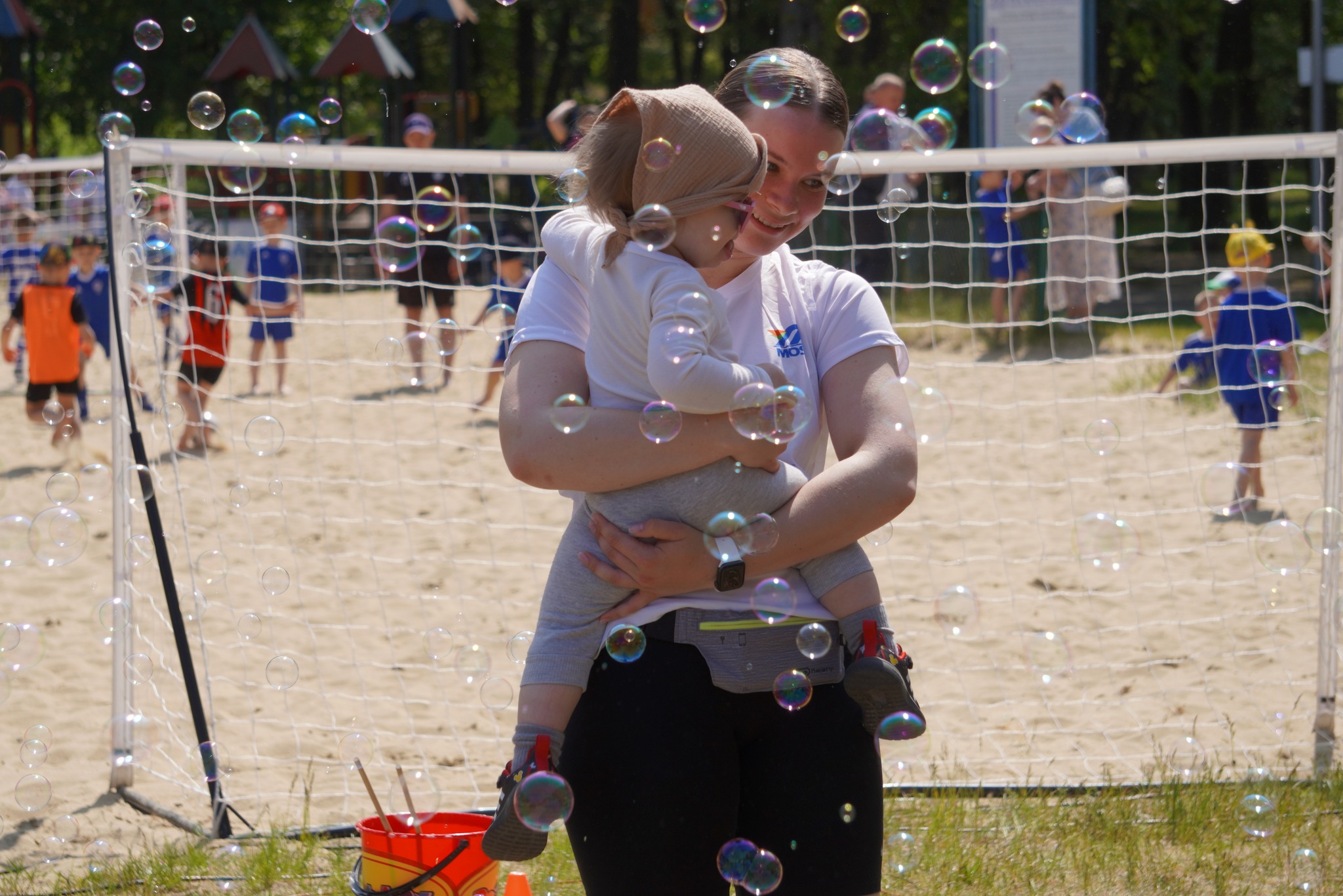 Kobieta z dzieckiem na ręku stoi na plaży wśród baniek mydlanych. W tle siatka i dzieci grające w piłkę.