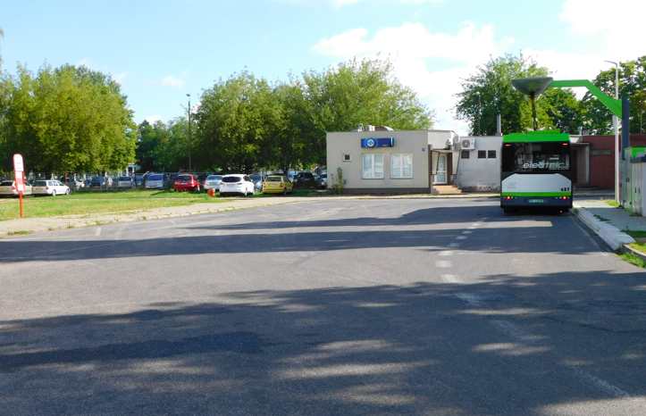 Utrudnienia na pętli przy ul. Kossaka Pętla autobusowa, po prawej stronie stacja ładowania i autobus elektryczny. Po lewej słupek z rozkładem jazdy, w oddali drzewa i zaparkowane auta osobowe.