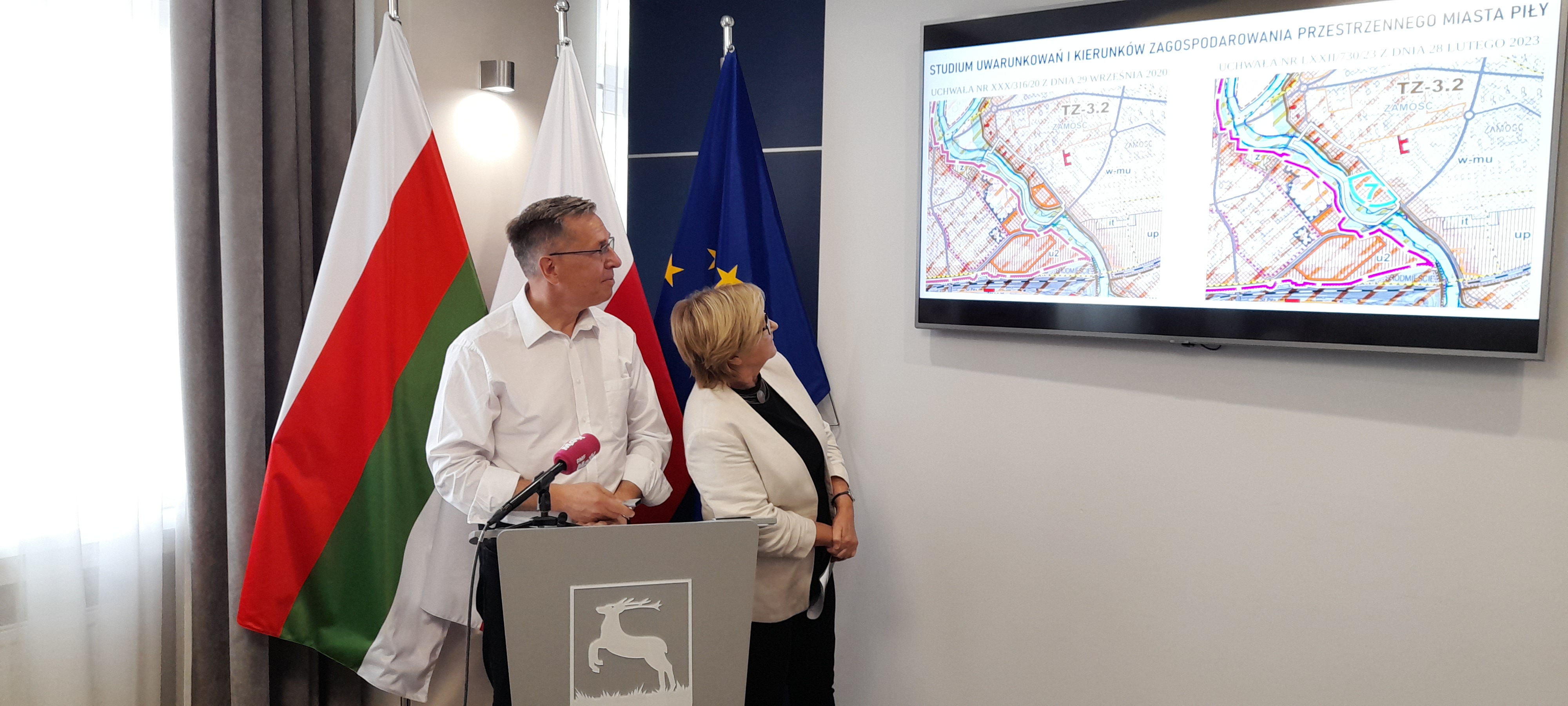 Prezentacja planu zagospodarowania przestrzennego miasta Piły w rejonie ulic Browarnej i Młodych. Kobieta i męczyna w reprezentacyjnym pomieszczeniu, stoją za mównicą i patrzą na plan wyświetlony na wisząym na ścianie mnitorze. W tle flagi: Piły, Polski i Unii Europejskiej.