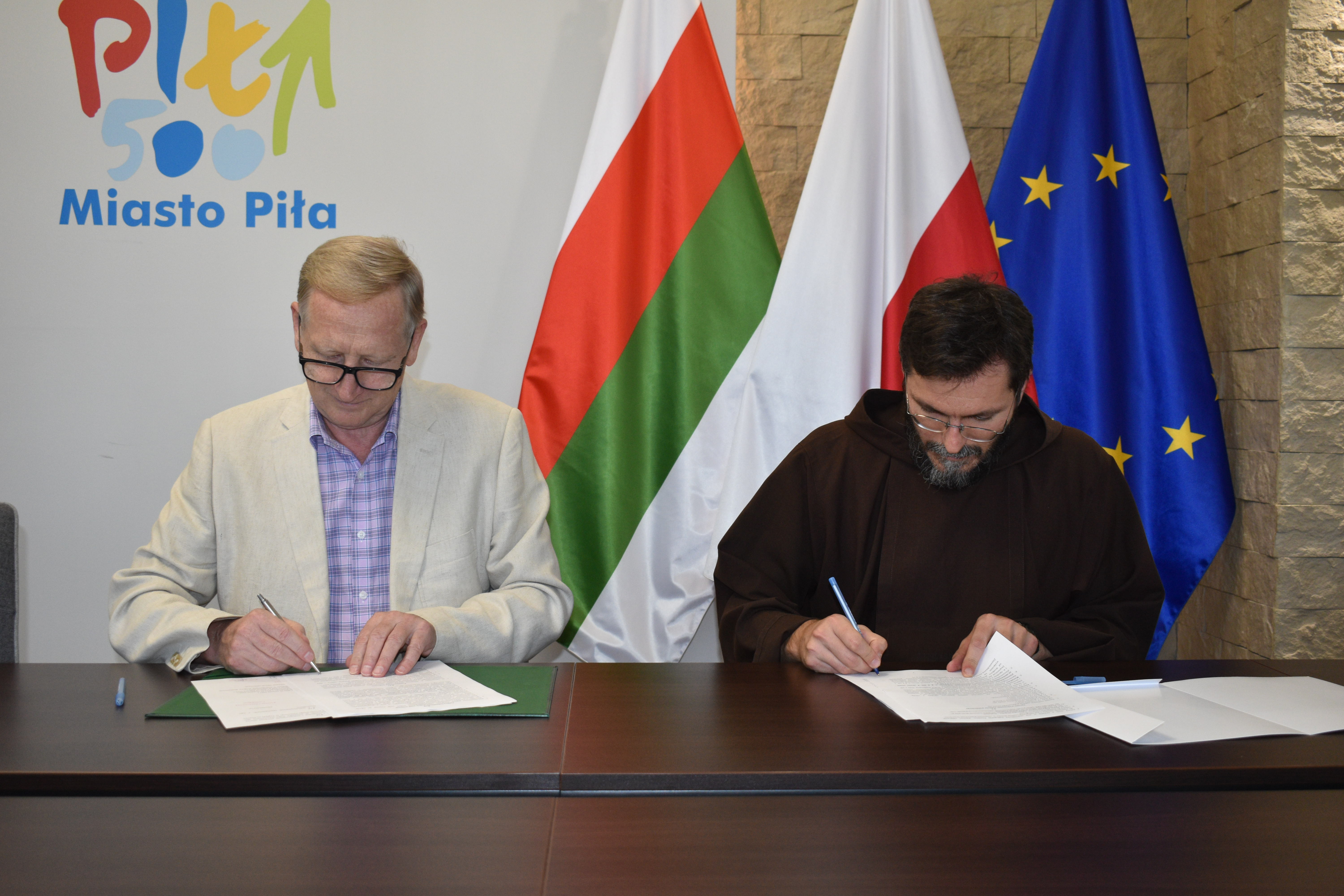 Mężczyzna w&nbsp;garniturze i&nbsp;duchowny w&nbsp;habicie siedzą za stołem w&nbsp;reprezentacyjnym pomieszczeniu i&nbsp;podpisują dokumenty. W tle logotyp i&nbsp;napis Piła Miasto oraz&nbsp;flagi: Piły, Polski i&nbsp;Unii Europejskiej.