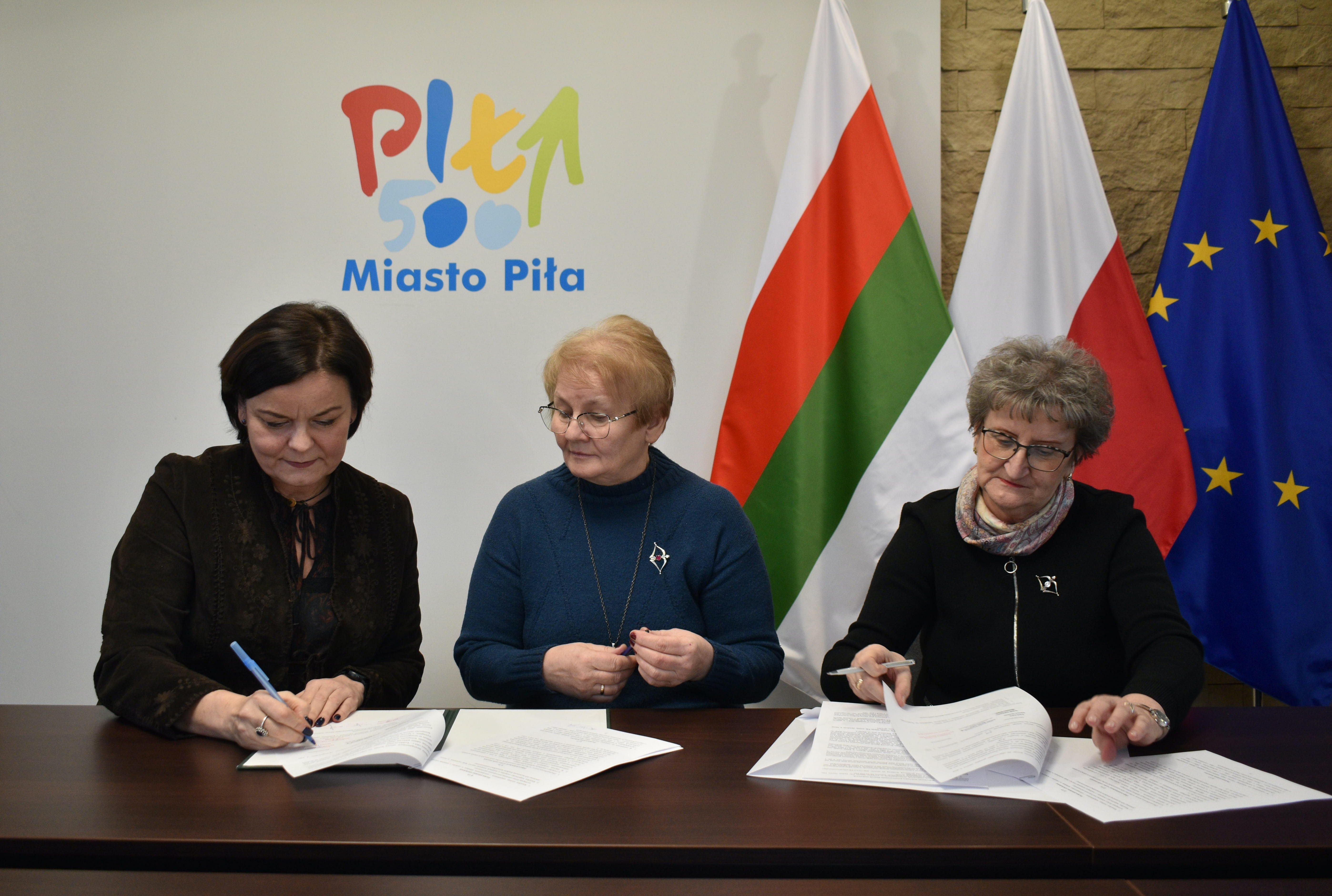 Trzy kobiety siedzące przy stole, podpisujące dokumenty. Za nimi flagi Piły, Polski i&nbsp;Unii Europejskiej.