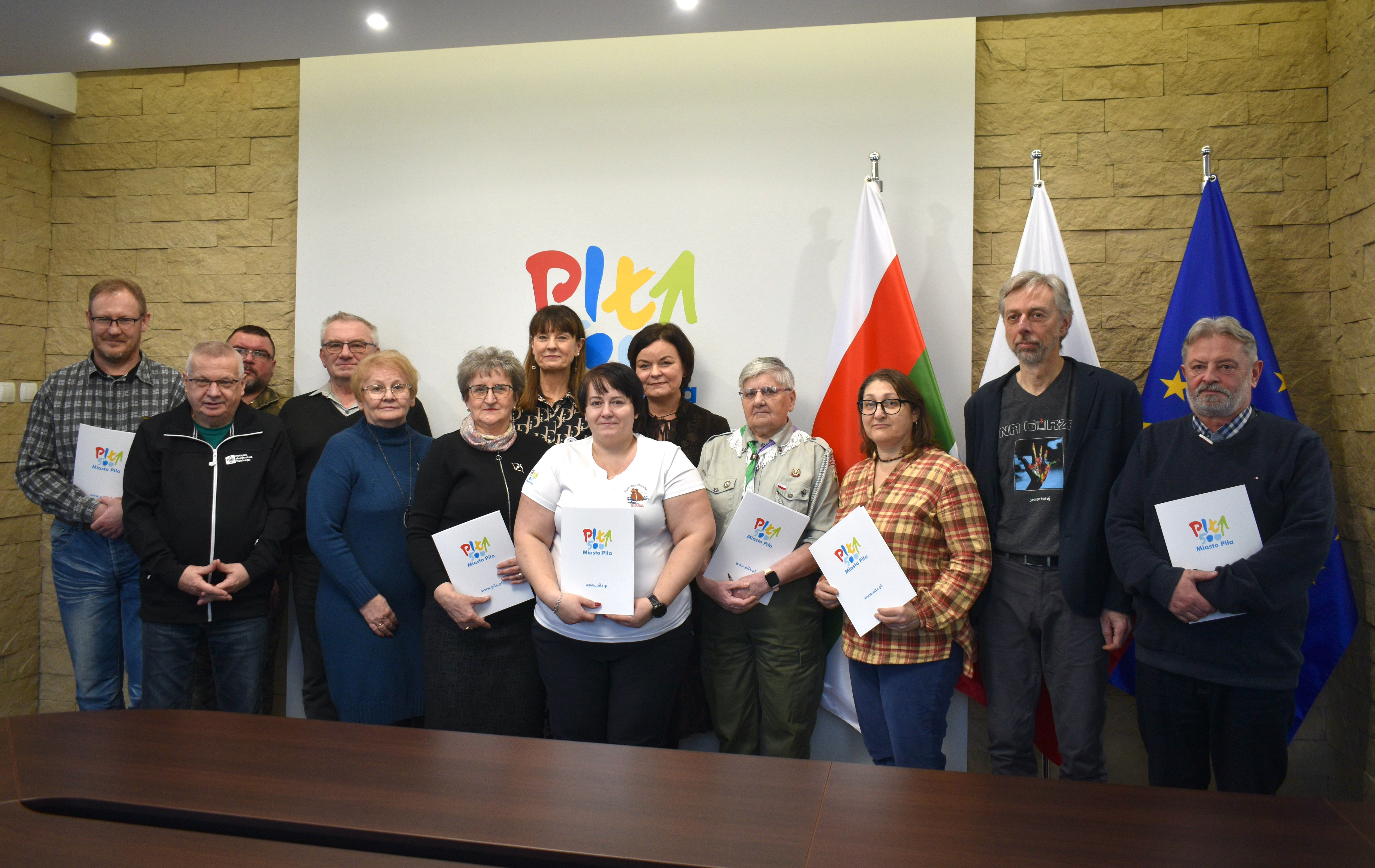 Grupa ludzi pozuje w&nbsp;pomieszczeniu, część z&nbsp;nich trzyma teczki z&nbsp;logotypem Miasto Piła.