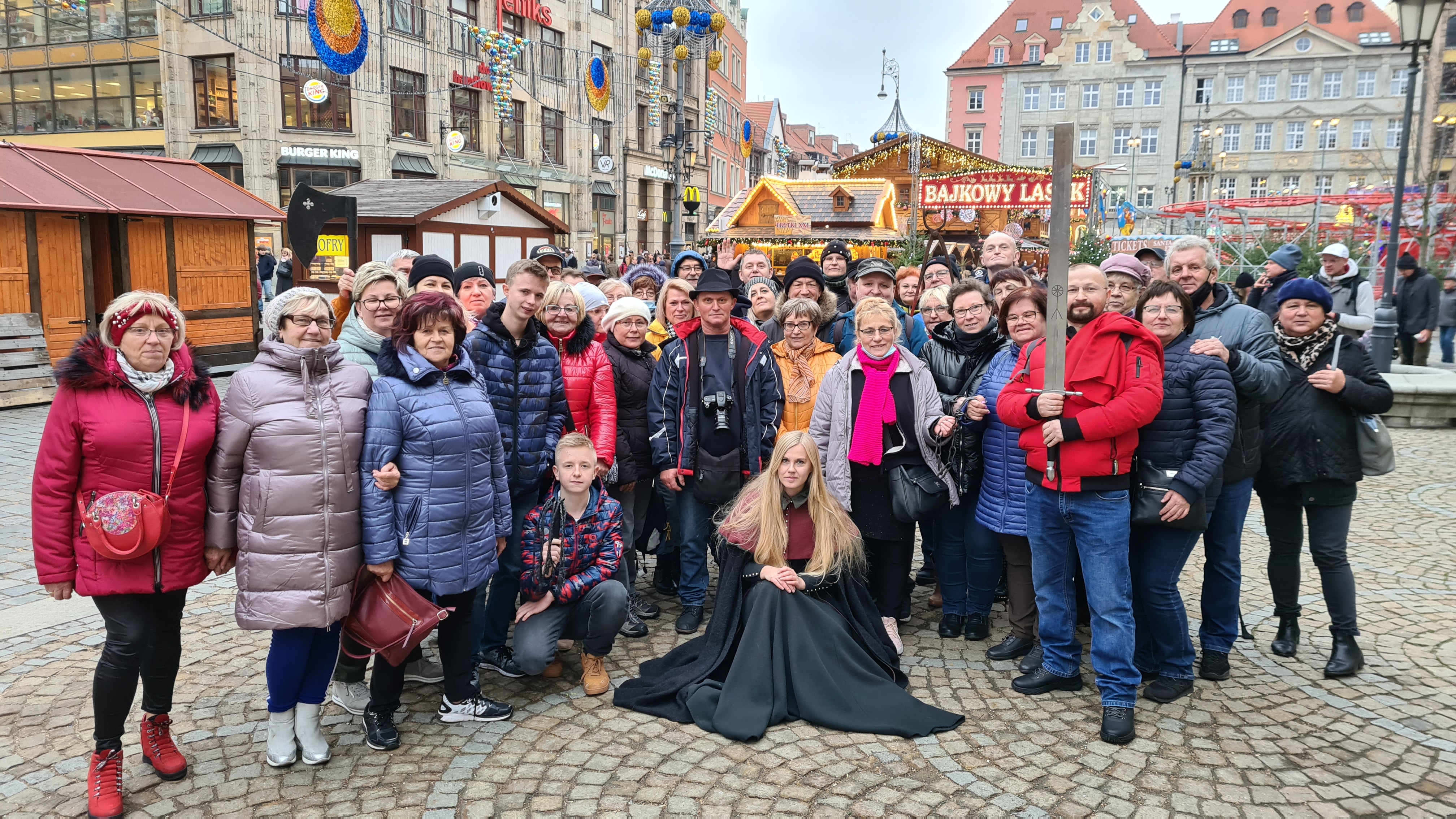 Grupa ludzi na&nbsp;świeżym powietrzu pozuje do zdjęcia, w&nbsp;tle jarmark miejski.