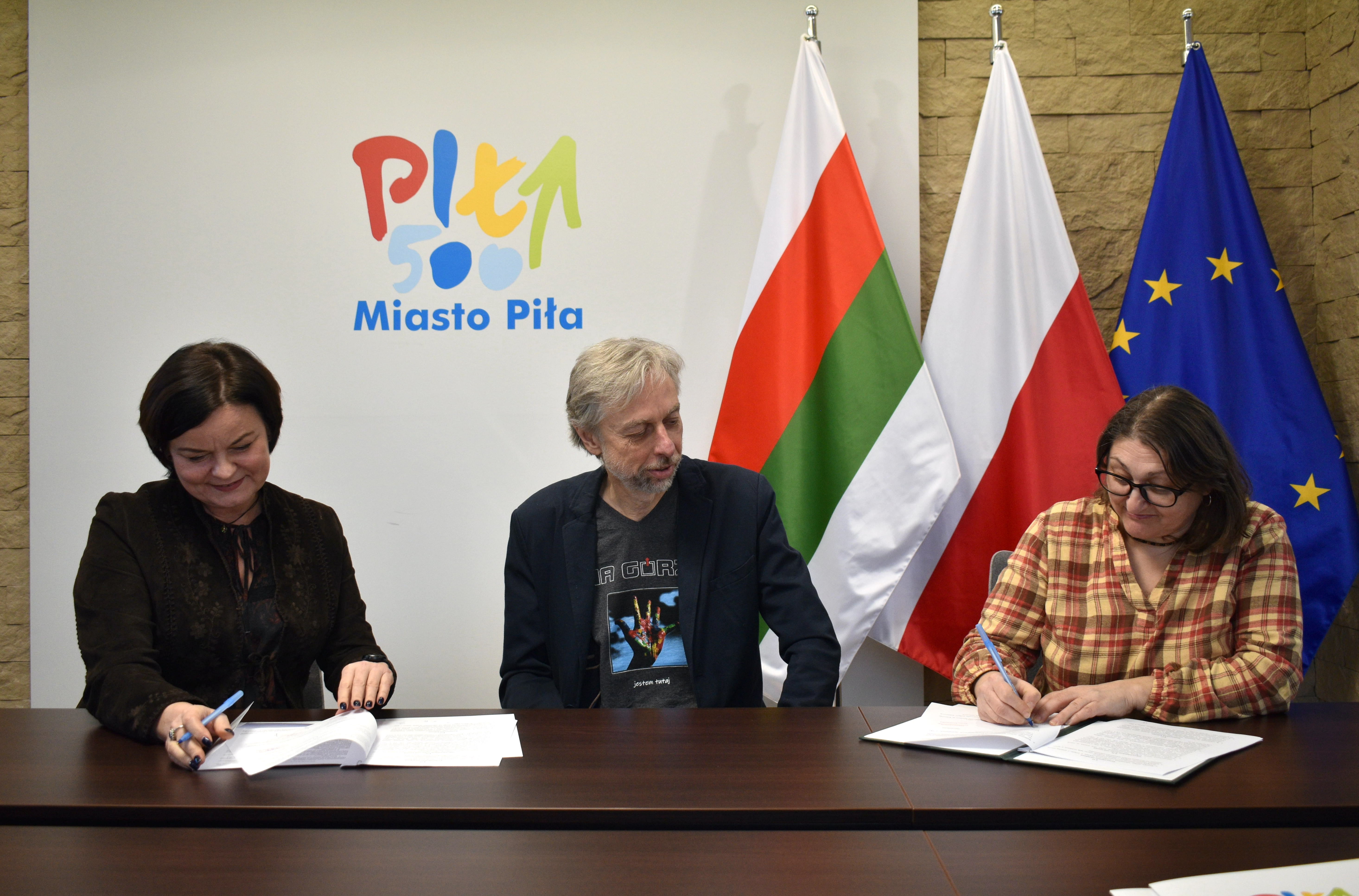 Mężczyzna i&nbsp;dwie kobiety siedzący przy stole w&nbsp;pomieszczeniu, podpisują dokumenty. Za nimi flagi Piły, Polski i&nbsp;Unii Europejskiej oraz&nbsp;logo Miasto Piła.