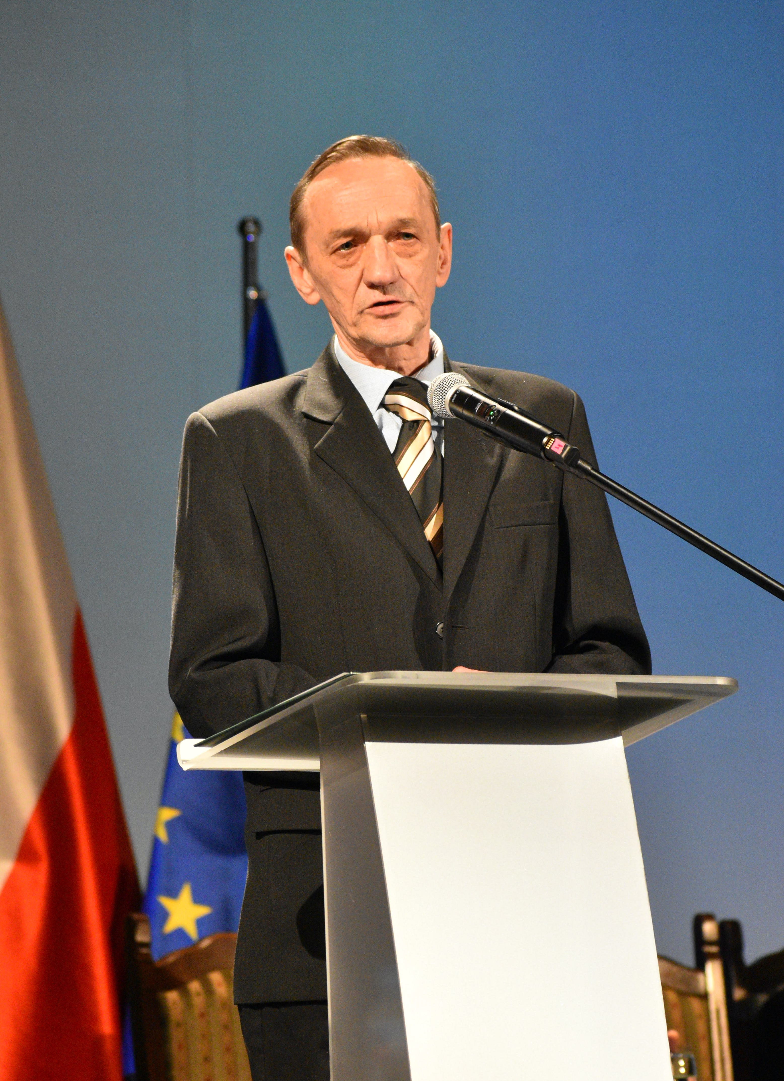 Dojrzały mężczyzna w&nbsp;garniturze stojący przy m&oacute;wnicy, wypowiada się do mikrofonu, w&nbsp;tle flagi