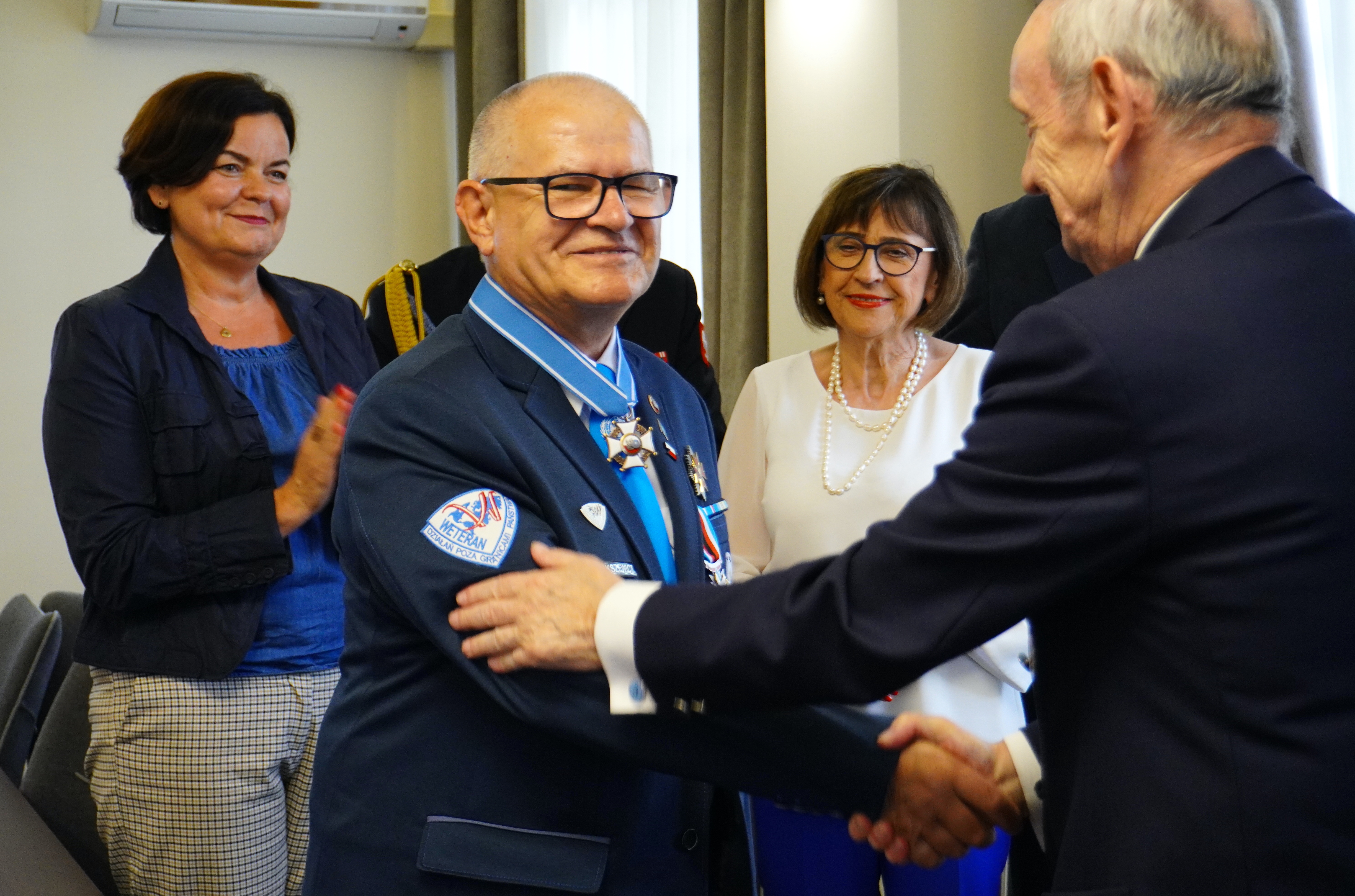 Na pierwszym planie dw&oacute;ch mężczyzn podaje sobie dłonie, jeden składa drugiemu gratulacje. Jeden z&nbsp;mężczyzn ma na&nbsp;ramieniu marynarki naszywkę weterana misji pokojowych ONZ, a&nbsp;na szyi medal - krzyż zasługi. W tle grupa ludzi klaszcze w&nbsp;dłonie, uśmiechają się. 