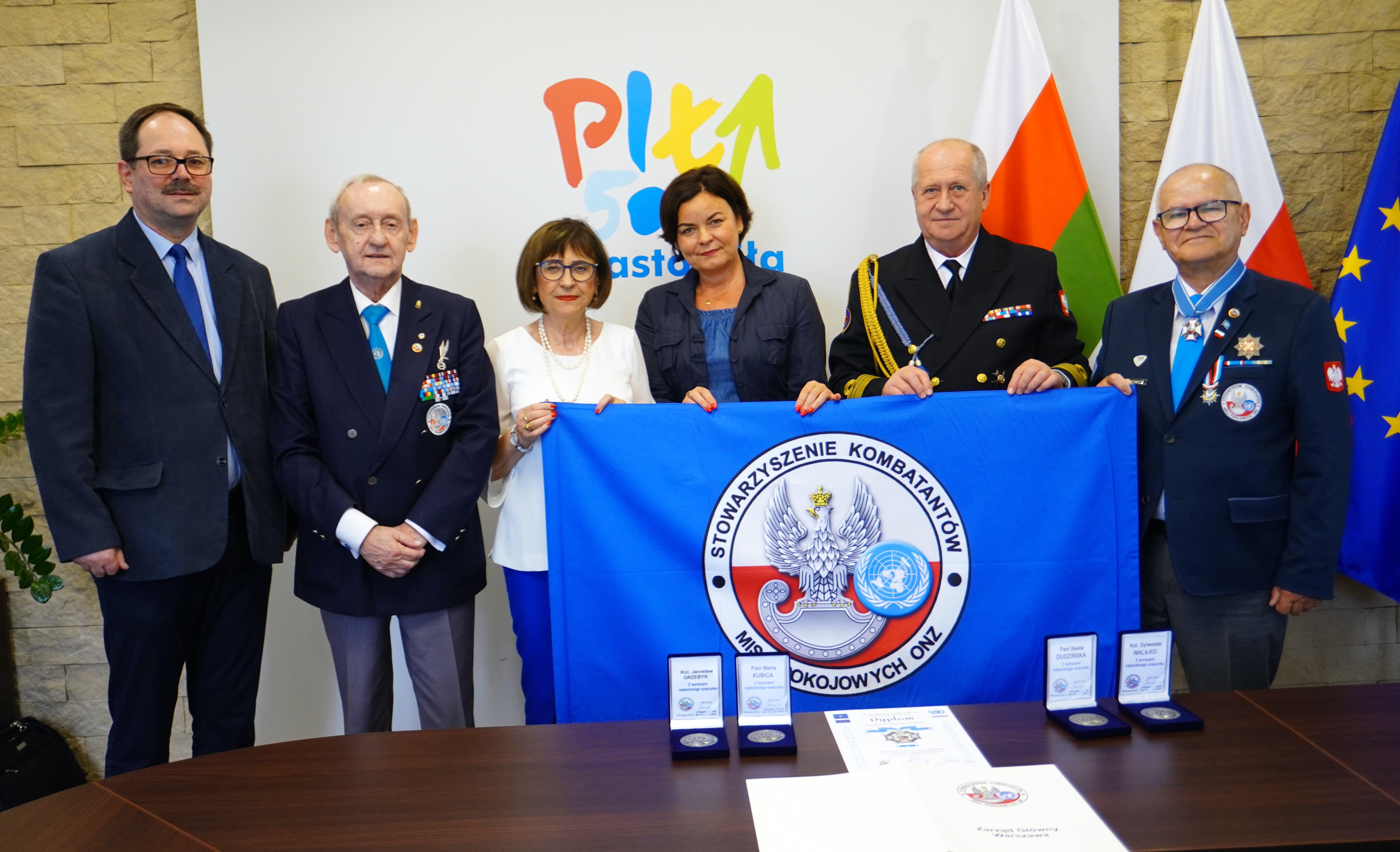 Grupa ludzi, dwie elegancko ubrane kobiety i&nbsp;czterech mężczyzn w&nbsp;mundurach, na&nbsp;kt&oacute;rych zawieszone są liczne odznaki. W pomieszczeniu stoją flagi miasta Piły, Polski i&nbsp;Unii Europejskiej. W tle na&nbsp;ścianie logo Miasto Piła. Ludzie trzymają flagę Stowarzyszenia Kombatant&oacute;w Misji Pokojowych ONZ, na&nbsp;stole przed nimi stoją pudełka z&nbsp;medalami i&nbsp;dyplom. 