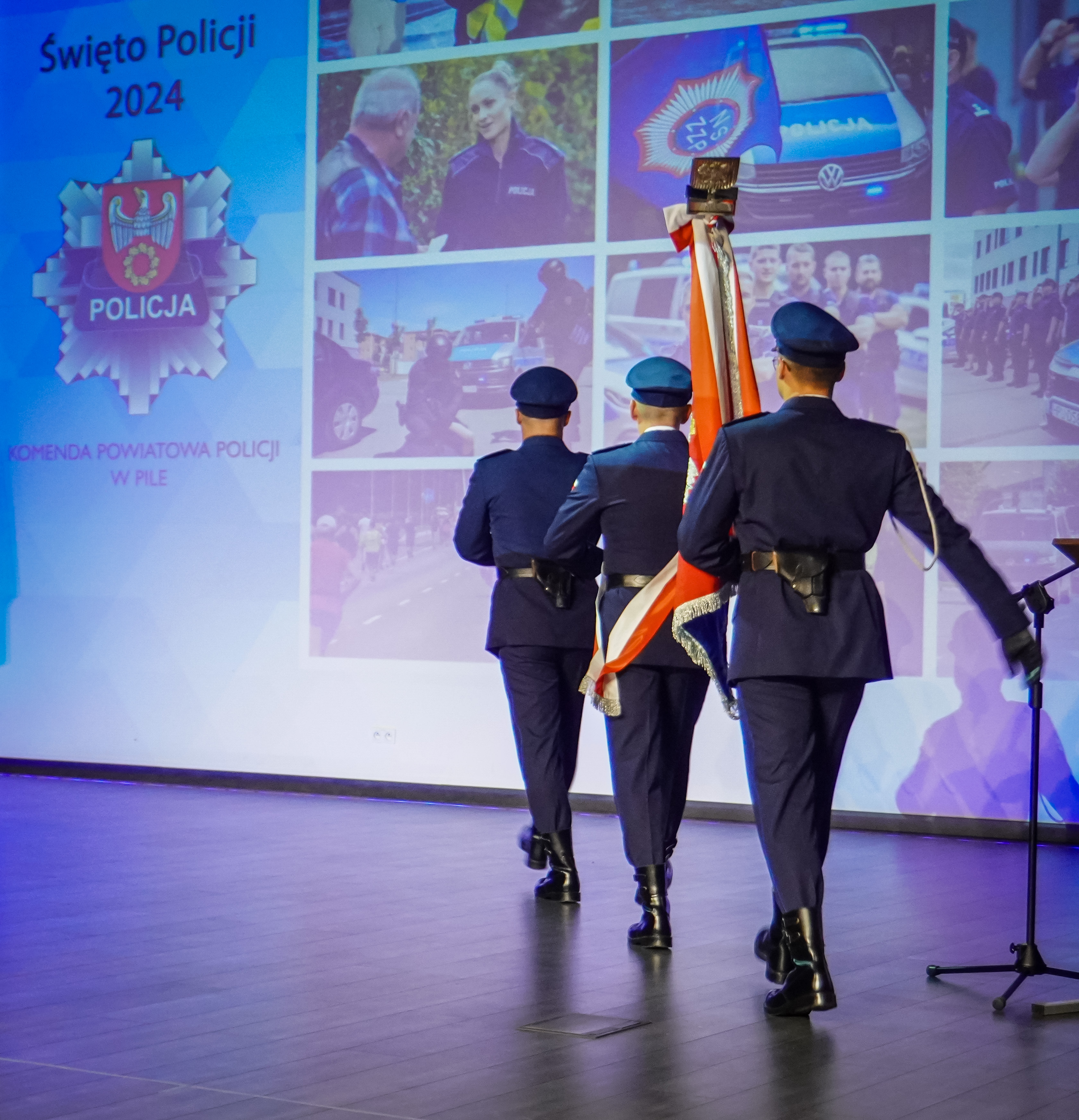 3 osoby w&nbsp;mundurach policyjnych niosący sztandar na&nbsp;scenie.