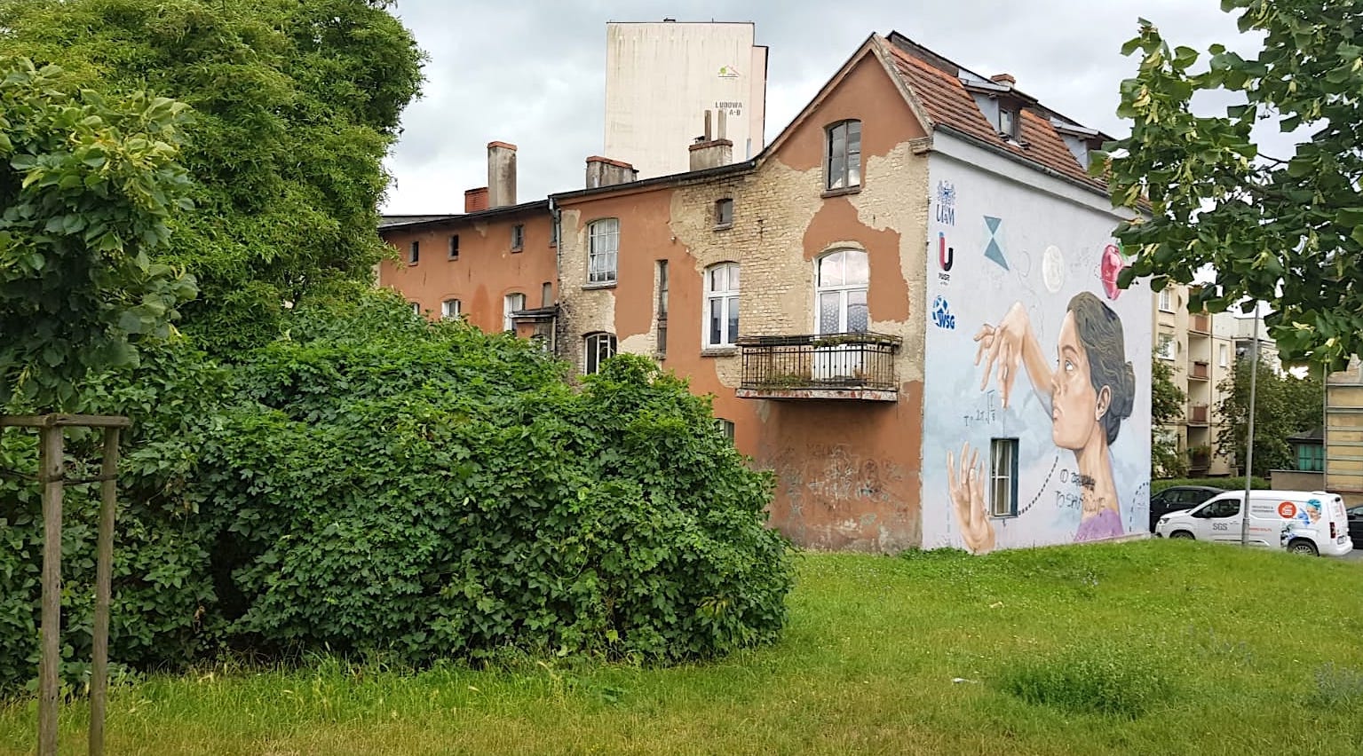 Stara kamienica, na&nbsp;jednej ze ścian mural przedstawiający twarz kobiety, ikony związane z&nbsp;nauką. Obok kamienicy drzewa, krzewy.