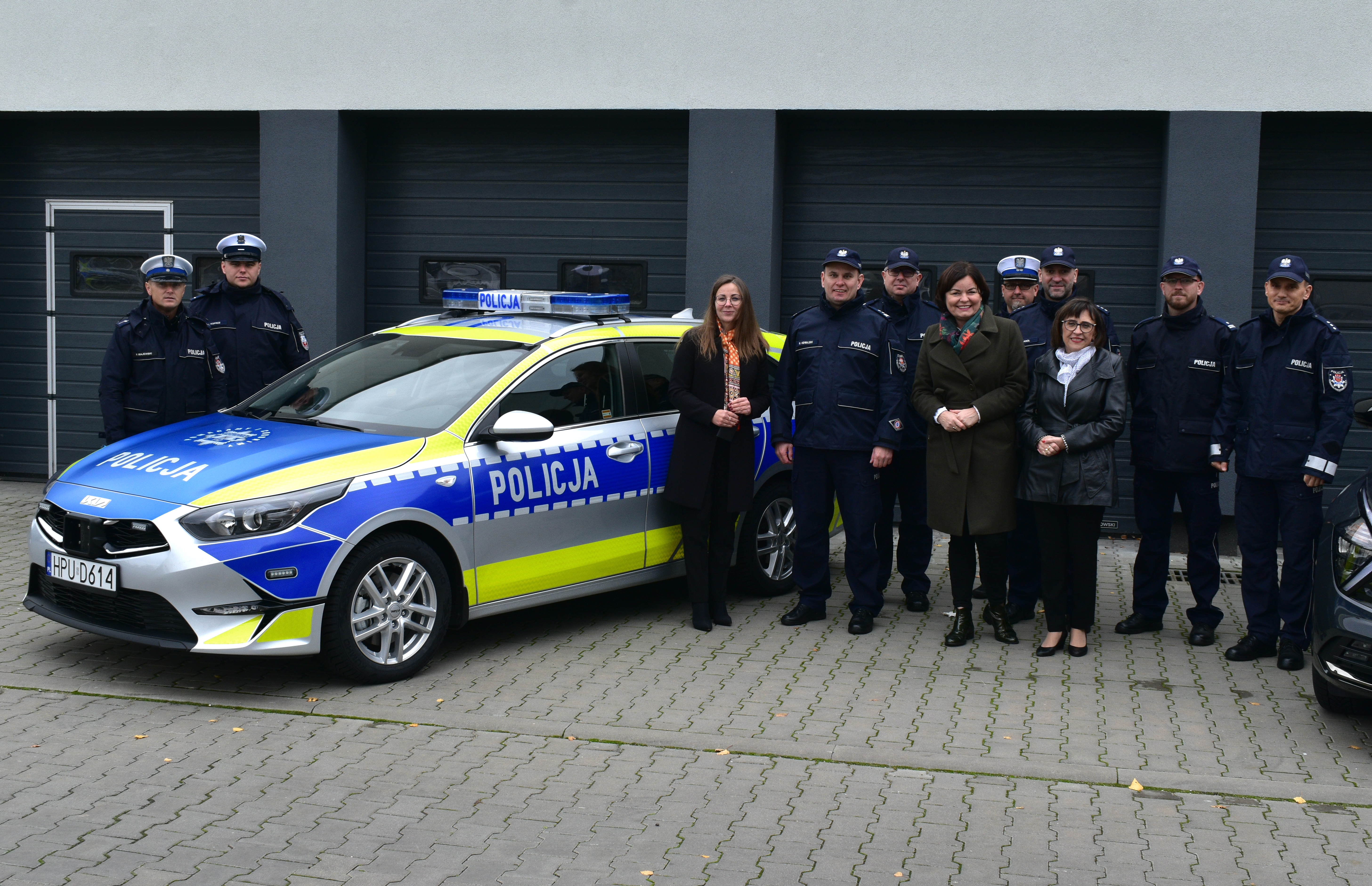 Grupa ludzi, w&nbsp;tym funkcjonariuszy policji stojących przed radiowozem