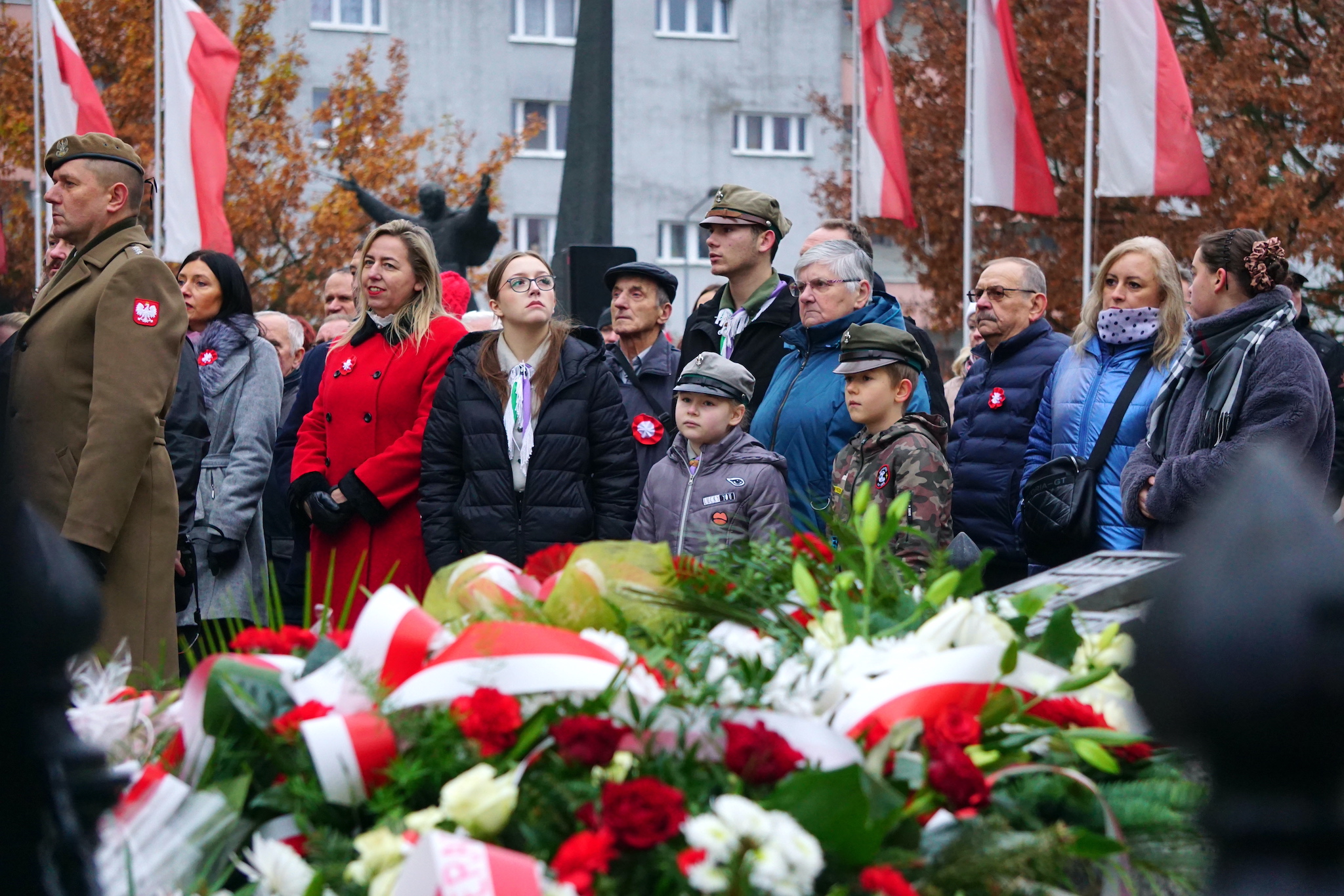 Grupa ludzi z&nbsp;biało-czerwonymi rozetami przypiętymi na&nbsp;piersi stoi na&nbsp;placu podczas uroczystości dnia niepodległości, na&nbsp;pierwszym planie złożone pod pomnikiem wieńce kwiat&oacute;w
